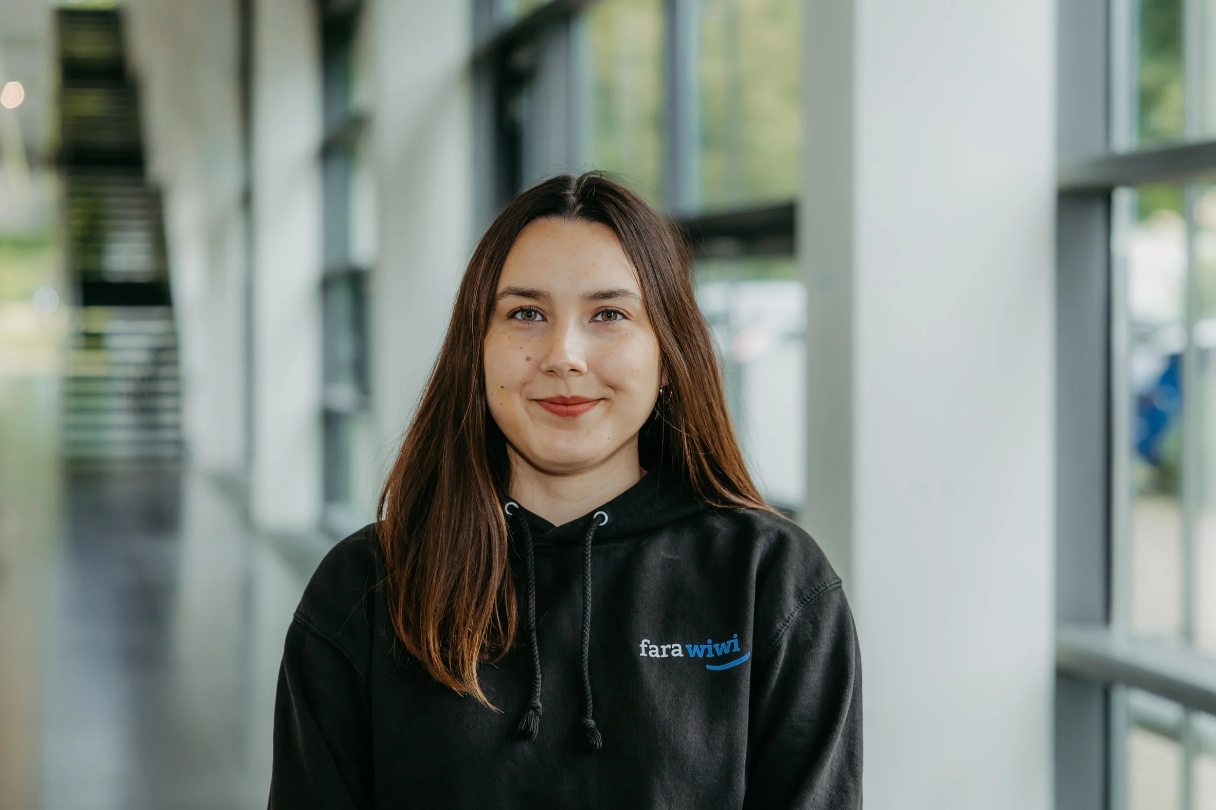 Junge Frau mit braunen Haaren und schwarzem Hoodie mit 'fara wiwi' logo, im Innenbereich neben großen Fenstern.