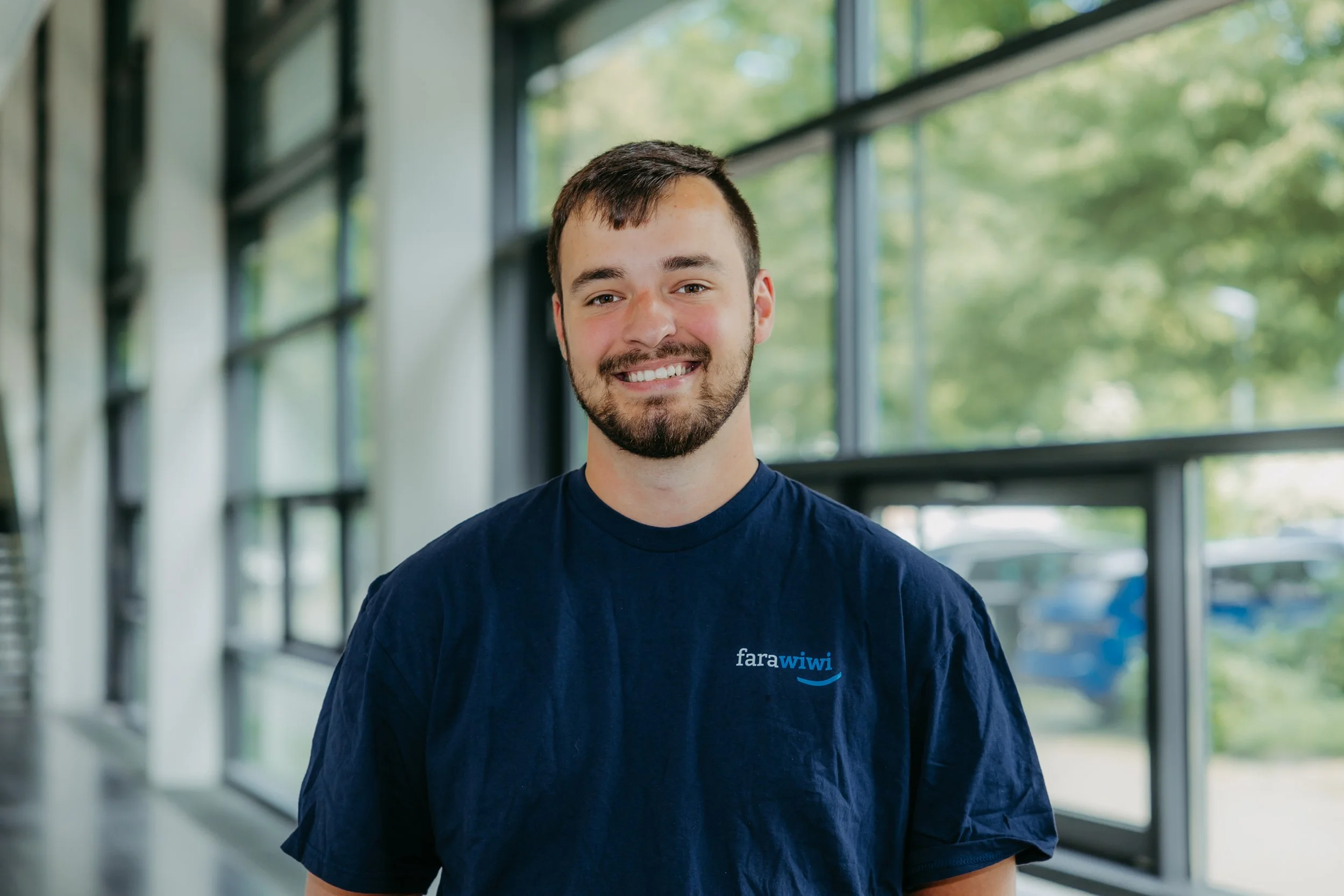 Ein lachender Mann mit dunklem Haar und Bart steht in einem hellen Raum mit großen Fenstern im Hintergrund, trägt ein dunkles T-Shirt mit dem Logo 'fara wiwi'.