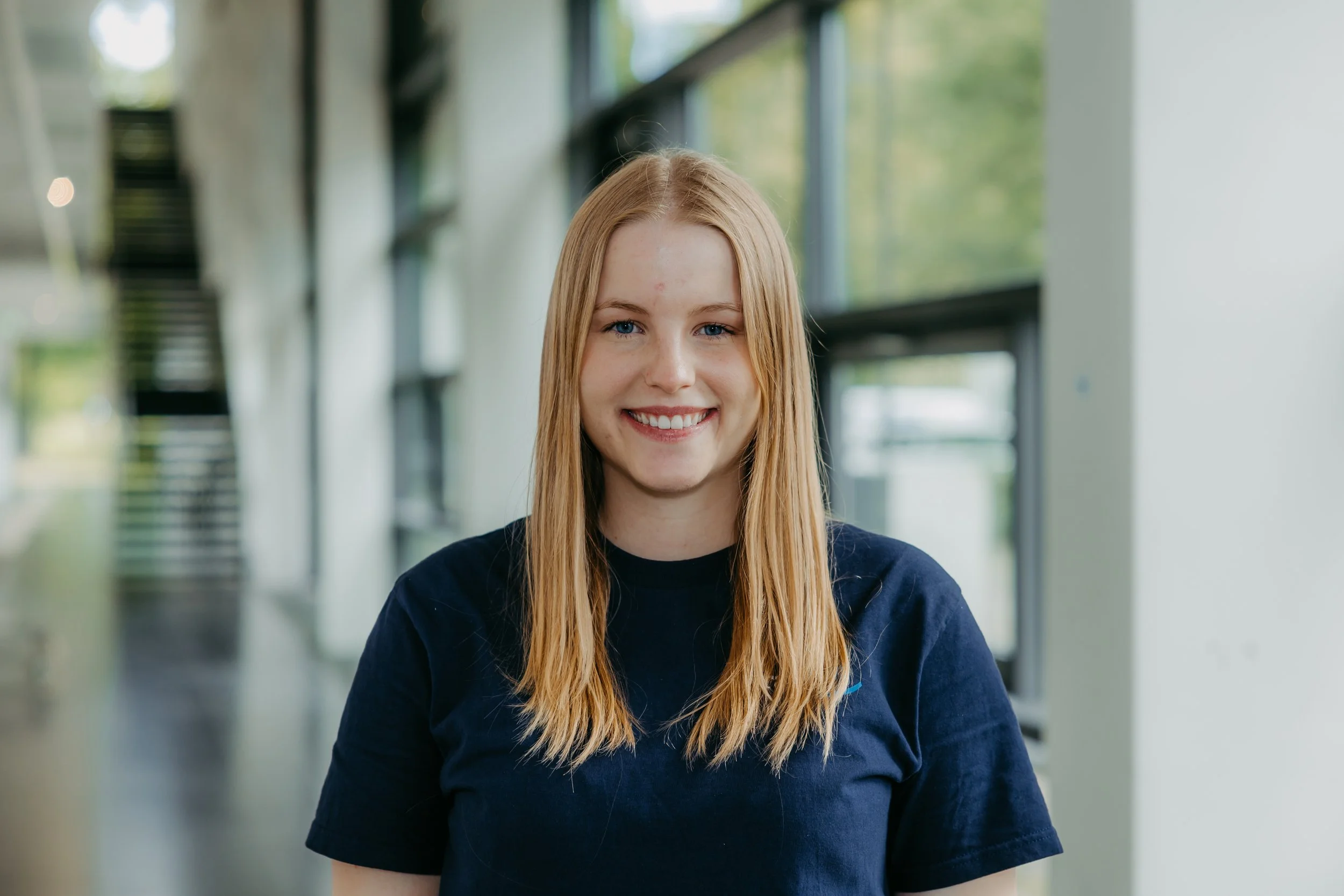 Junge Frau mit longitudinalen blonden Haaren, lächelnd, im Hintergrund ein moderner Flur mit großen Fenstern und Treppe