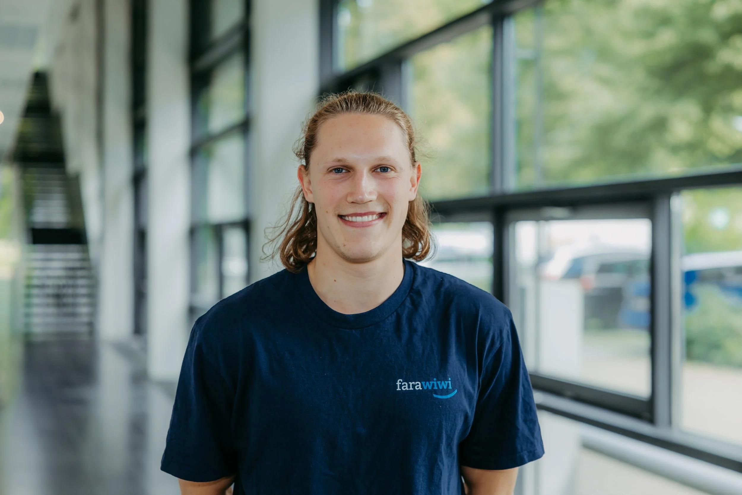 Ein junger Mann mit langen Haaren und einem blauen T-Shirt mit dem Logo "fara wiwi" in einem modernen Gebäude mit großen Fenstern und grünen Bäumen im Hintergrund.