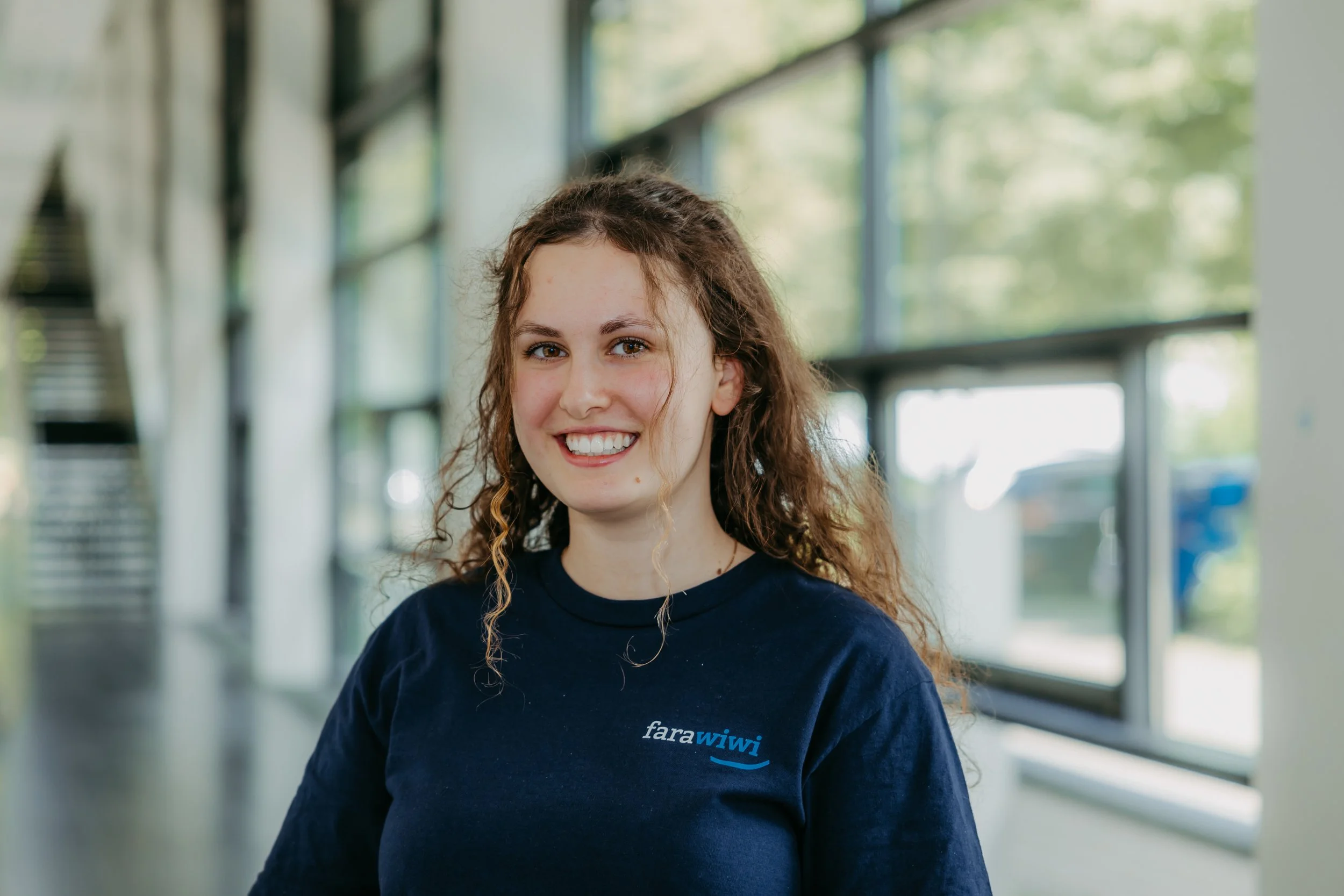 Junge Frau mit braunen, lockigen Haaren, trägt ein dunkelblaues T-Shirt mit dem Logo 'farawiwi', steht in einem hell erleuchteten Raum mit großen Fenstern im Hintergrund.