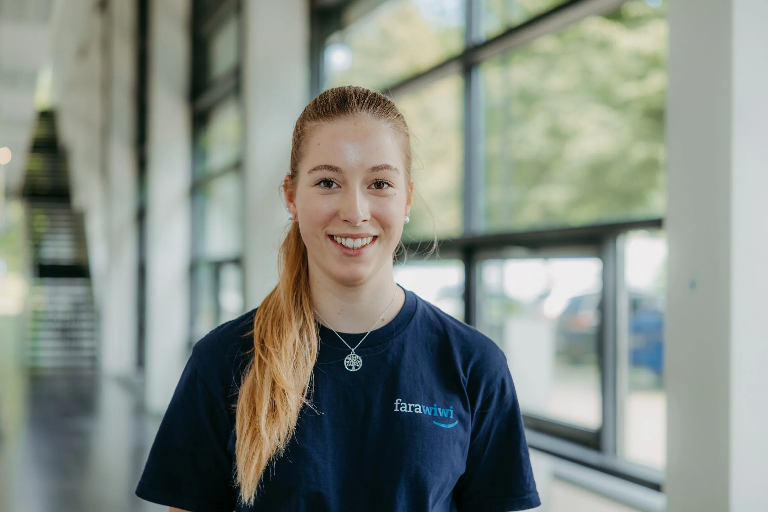 Lächelnde junge Frau mit roten Haaren, die eine dunkelblaue T-Shirt trägt mit Aufdruck 'farawiwi', im Hintergrund eine moderne, helle Raum mit großen Fenstern und grüner Landschaft draußen.