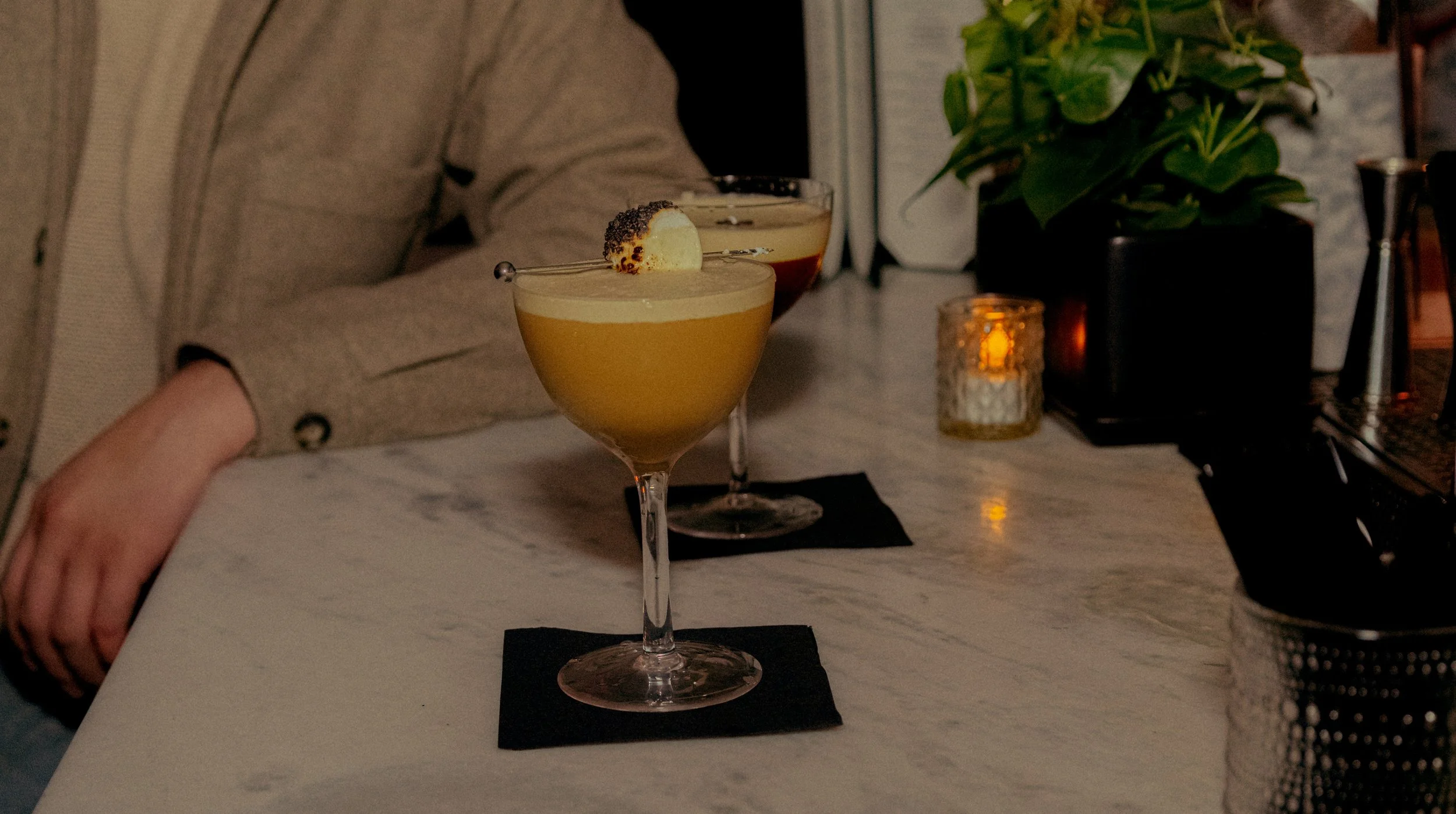 Two cocktails on a white marble bar counter with a dark background. The cocktails are garnished, one with a slice of citrus and black pepper, the other with dark berries. A person's arm is partially visible on the left, and a potted plant and a lit c