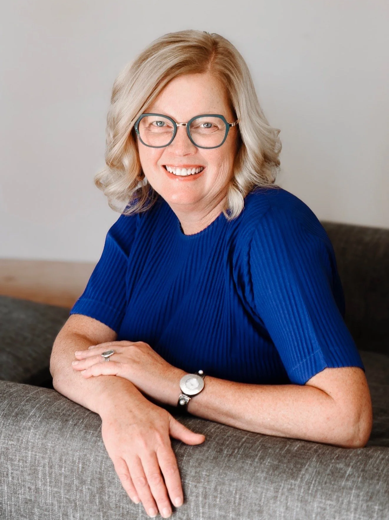 A smiling middle-aged woman with blonde hair, glasses, wearing a royal blue top, sitting on a gray sofa with her arms crossed on the armrest.