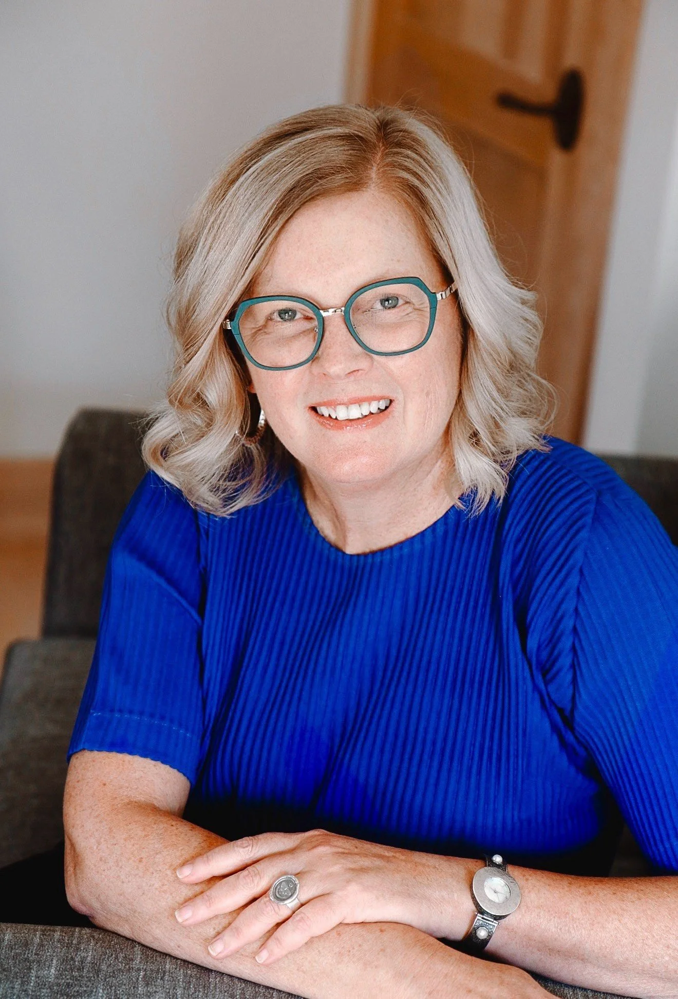A woman with blonde hair, wearing glasses and a bright blue top, smiling while sitting at a table indoors.