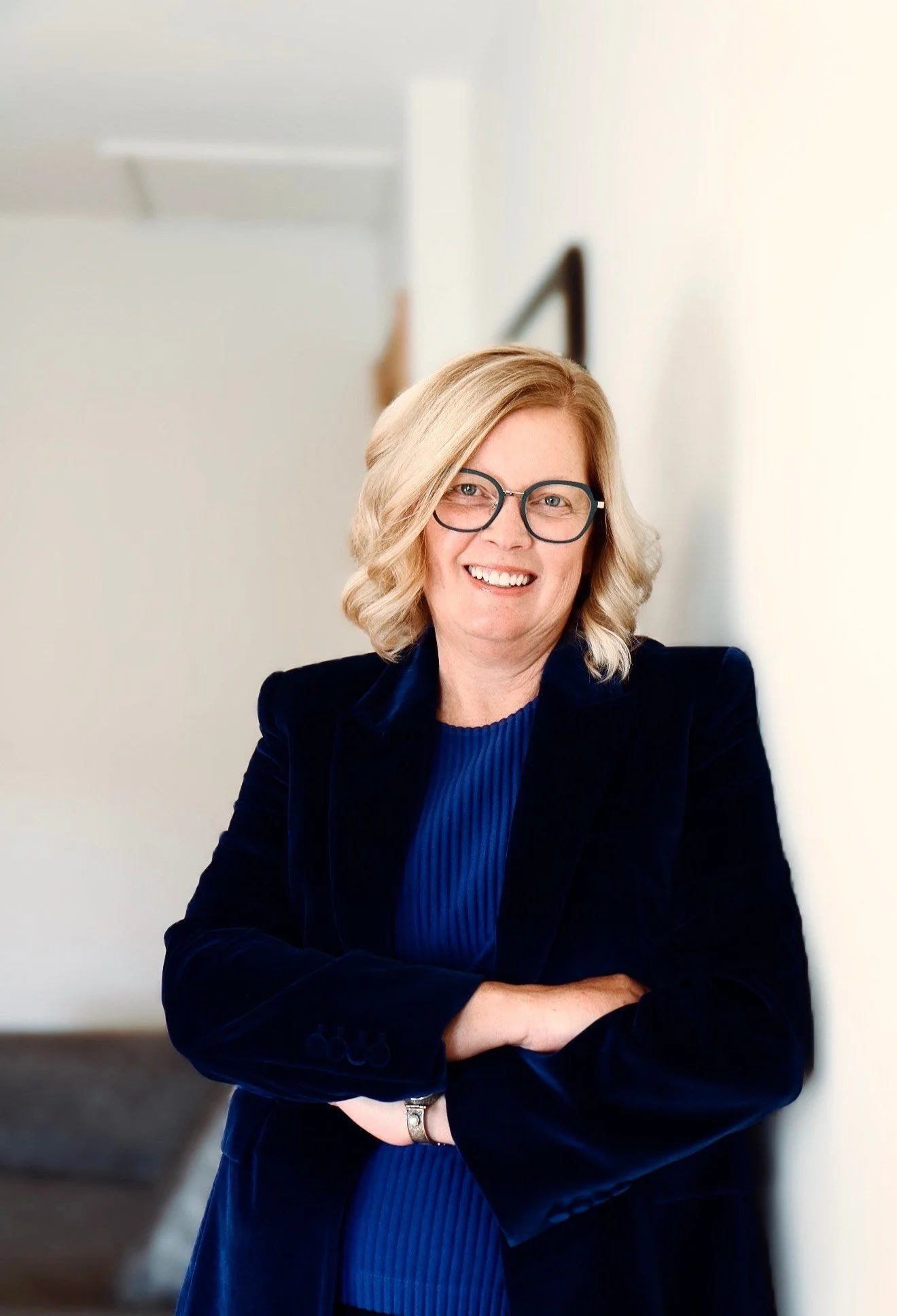 Woman with blonde hair and glasses smiling, standing with her arms crossed in a professional setting.