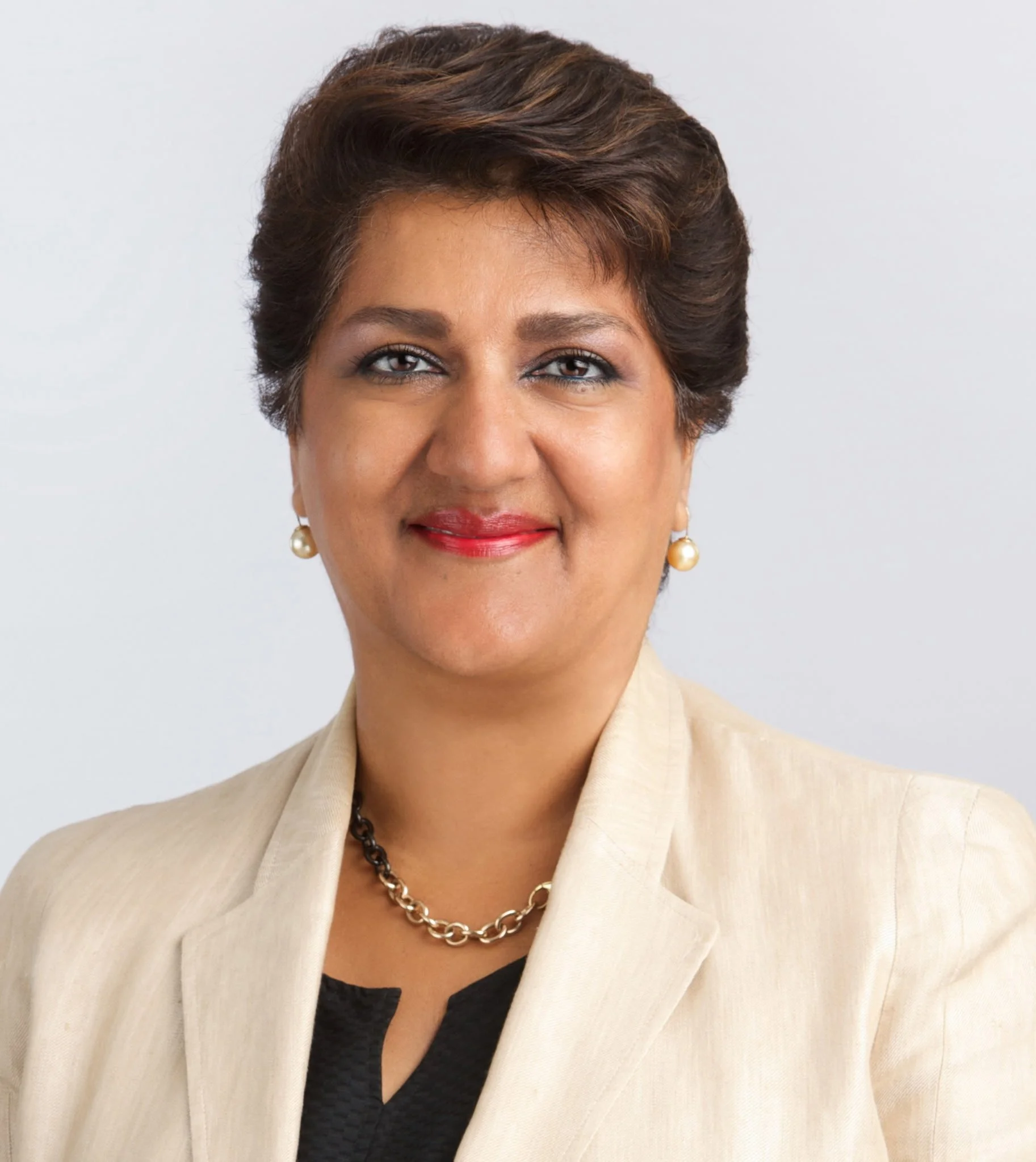 A professional woman with short hair, wearing pearl earrings, a necklace, a beige blazer, and a black top, smiling at the camera.