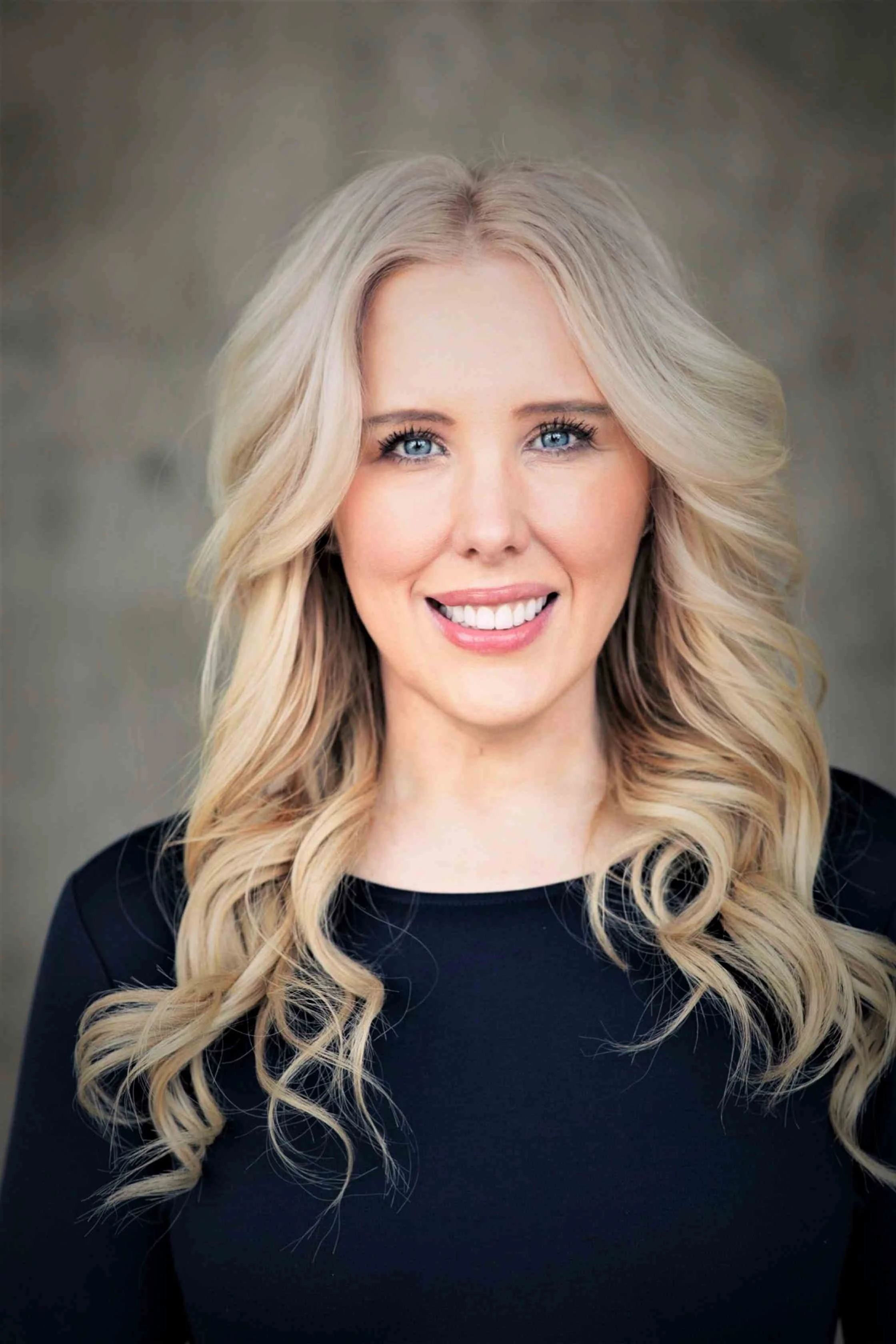 A woman with long, wavy blonde hair, blue eyes, and fair skin, smiling and wearing a black top, against a neutral background.