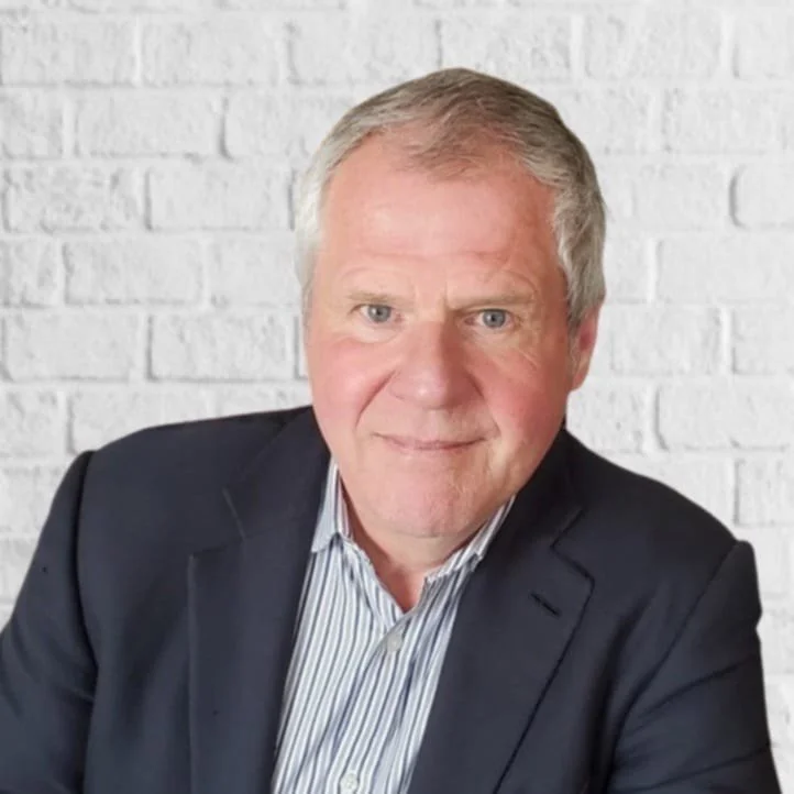 A middle-aged man with light hair wearing a dark blazer and a striped shirt, smiling with a white brick wall in the background.