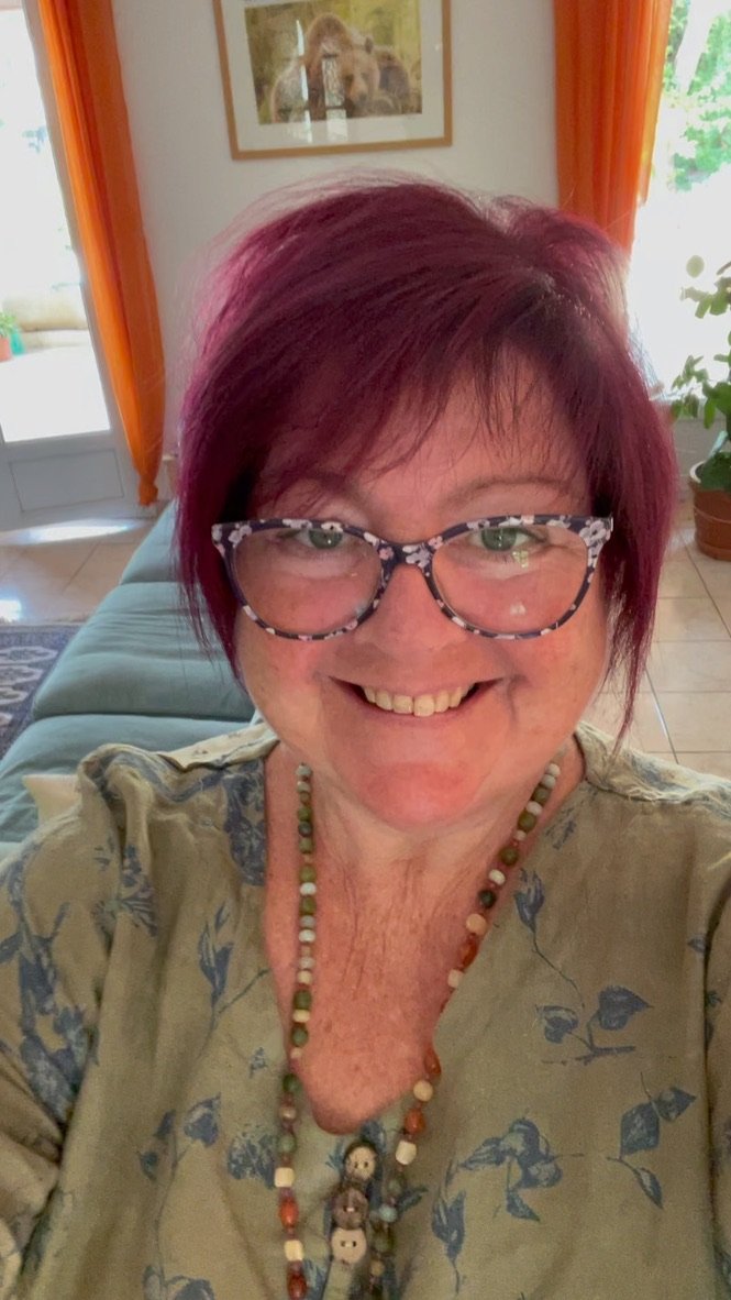 A smiling woman with short, burgundy hair, wearing glasses with a patterned frame, a floral blouse, and a beaded necklace. She is indoors with a window, orange curtains, and artwork on the wall behind her.