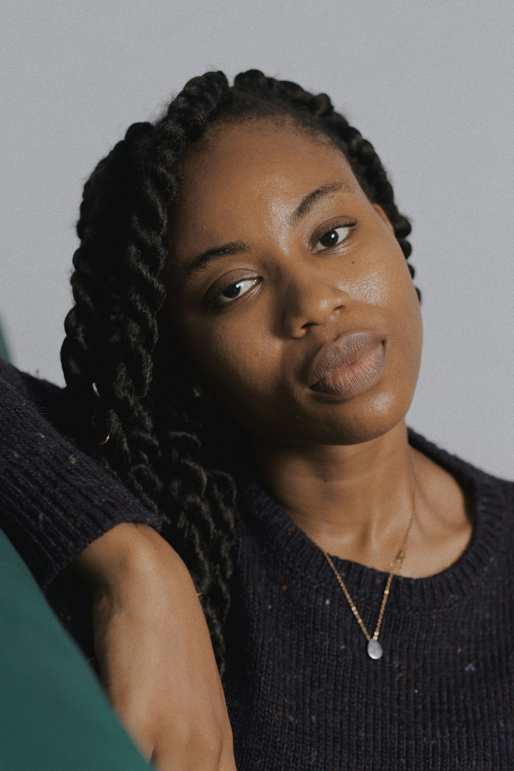 A close-up of a woman with dark brown skin and textured, twisted hair. She is wearing a dark, textured top and layered gold necklaces. Her expression is calm, and she is leaning slightly to one side, with her arm extended in front of her.