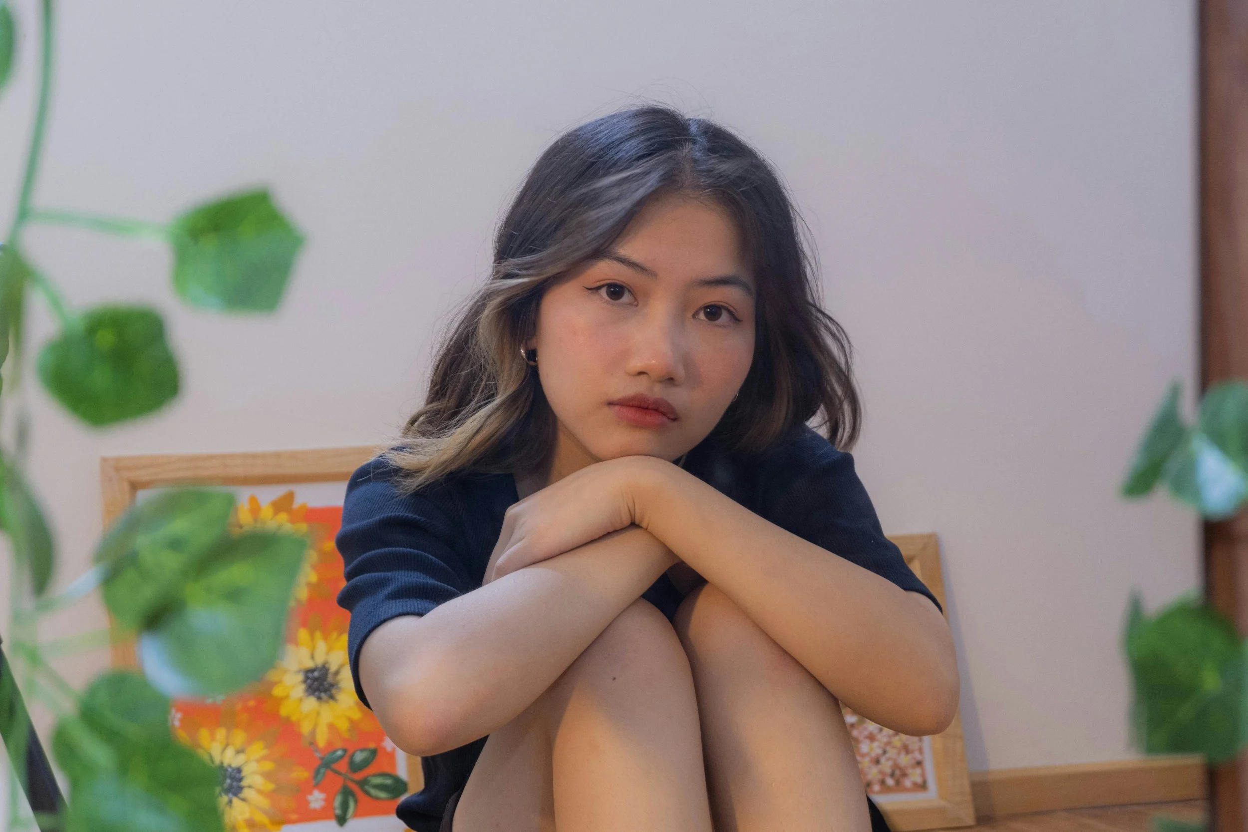 Portrait of a young woman with wavy hair and a contemplative expression, sitting indoors with her knees drawn up, surrounded by blurred green leaves in the foreground and colorful wall art in the background.