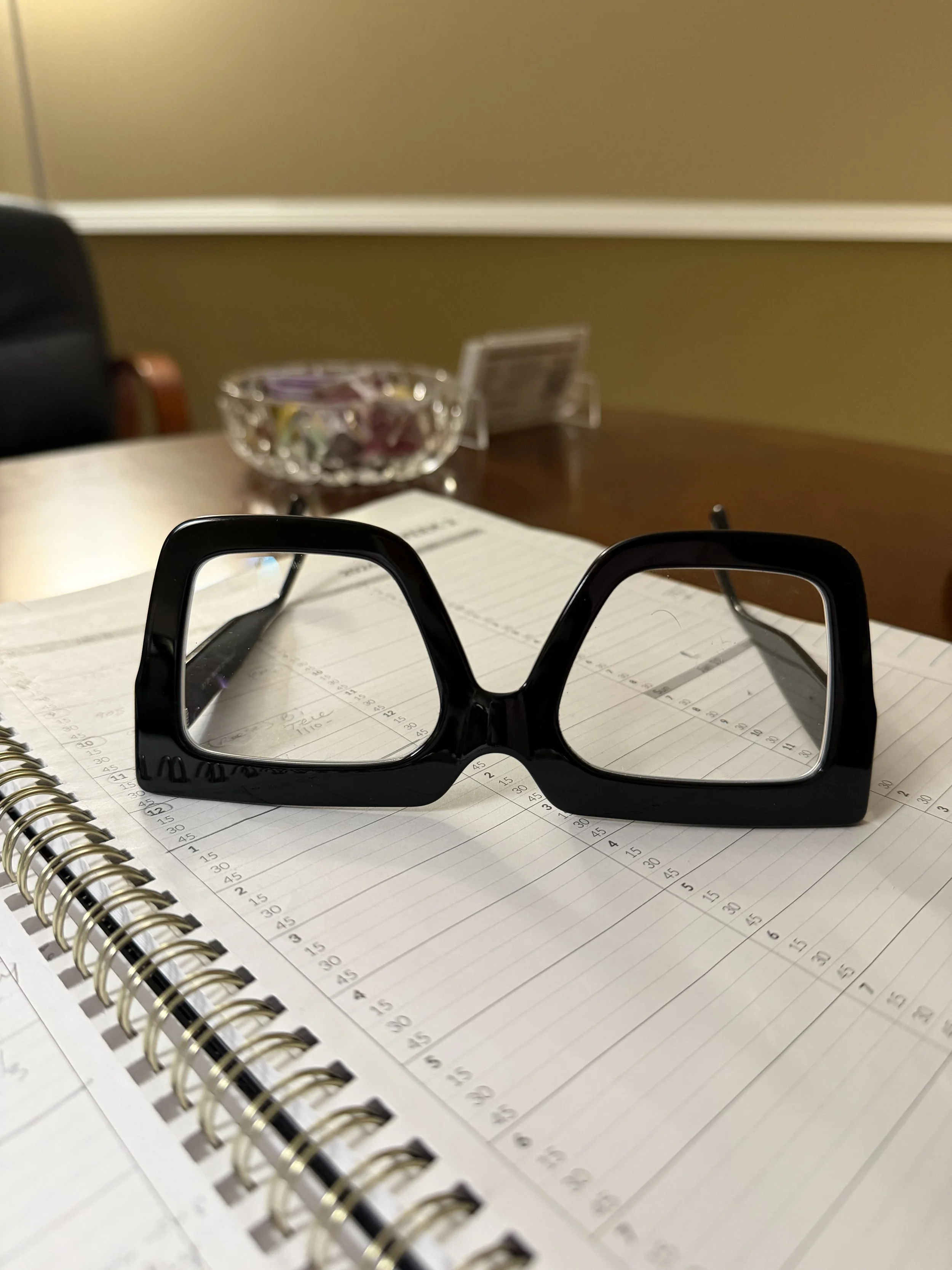 A pair of black glasses on an open spiral notebook filled with handwritten notes, with a computer screen, a crystal bowl, and a small sign or card in the blurry background.
