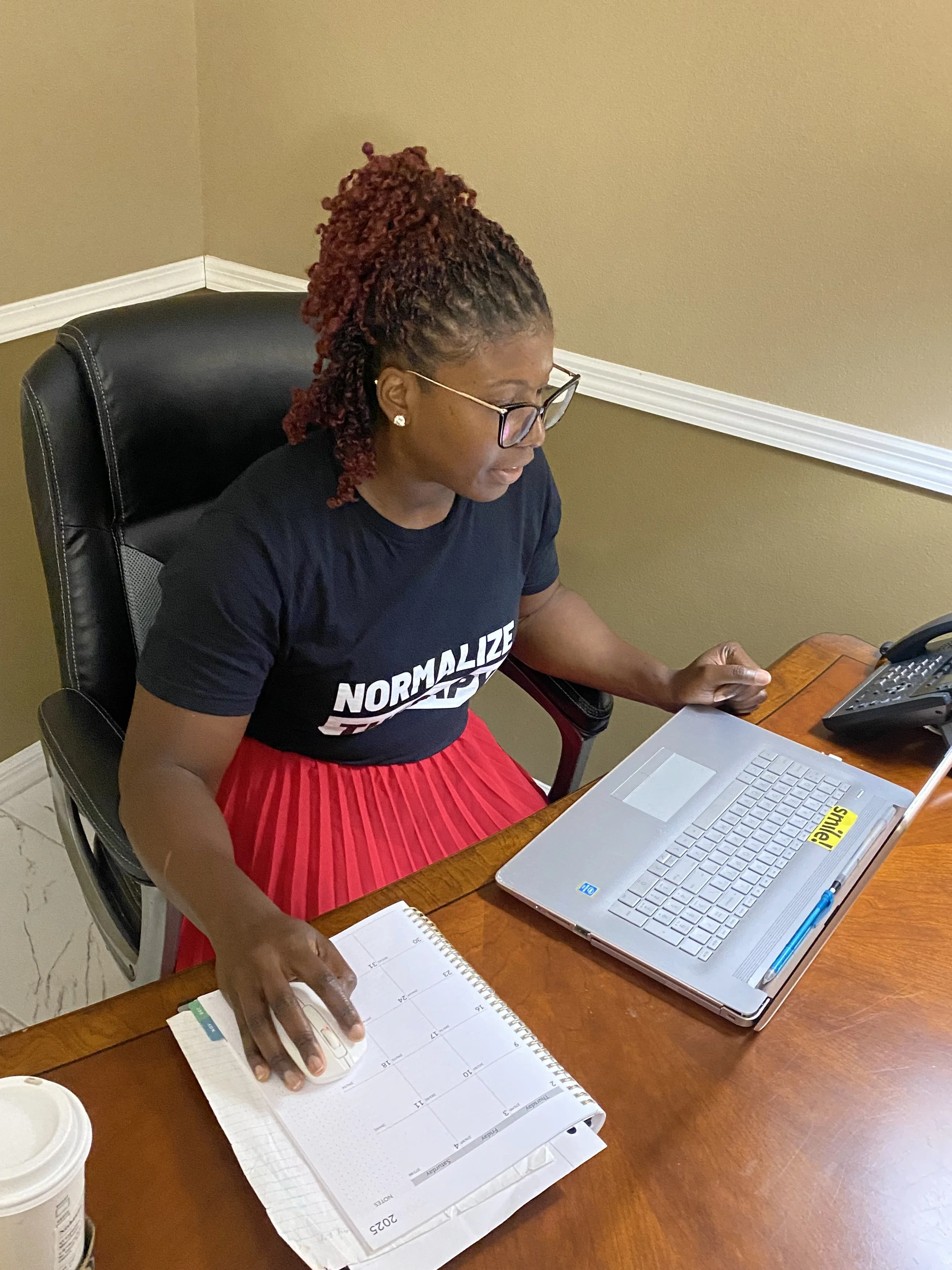 A woman with glasses and curly red hair, wearing a black T-shirt with the message 'Normalize T...,' sits at a wooden desk with a laptop, a calendar planner, a notebook, and a computer mouse. She appears to be working or planning, with a cup of coffee or tea nearby.