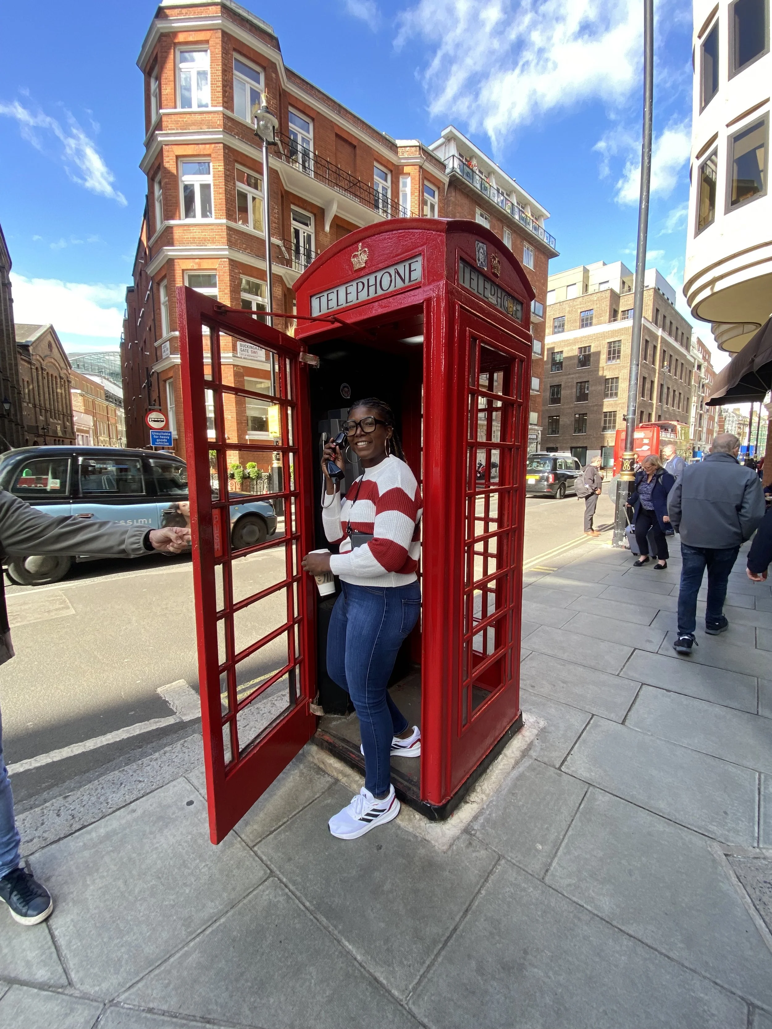 A grief therapist in a red and white striped sweater standing inside a classic red British telephone booth on a busy city street with buildings, cars, and pedestrians.