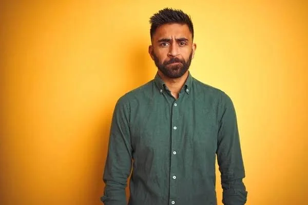 A man with dark hair and beard wearing a green button-up shirt stands against a yellow wall.