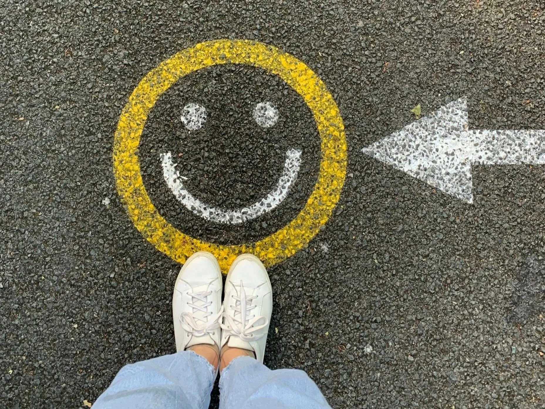 Person wearing white sneakers and light-colored jeans standing on asphalt with a smiley face and a yellow circle around it, pointing to the right.