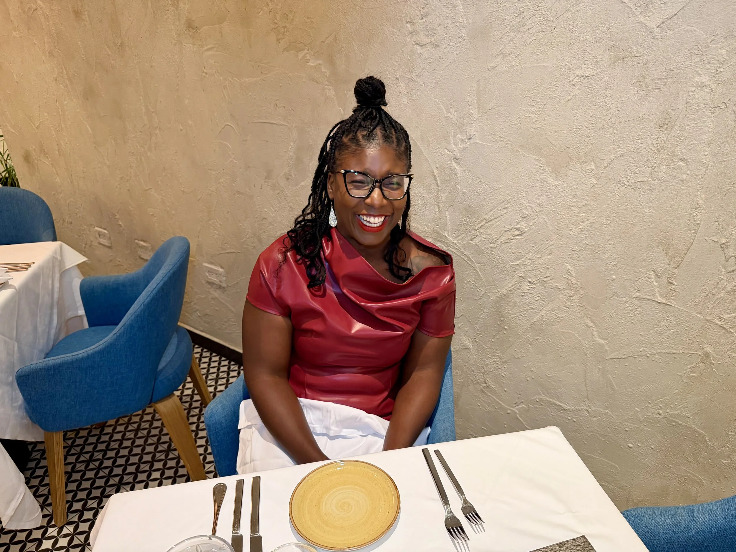A woman with glasses and long braids tied in a bun, wearing a red satin top and white pants, sitting at a restaurant table, smiling brightly. The table has a yellow plate, silverware, and a napkin. The background features a textured beige wall and additional blue chairs.