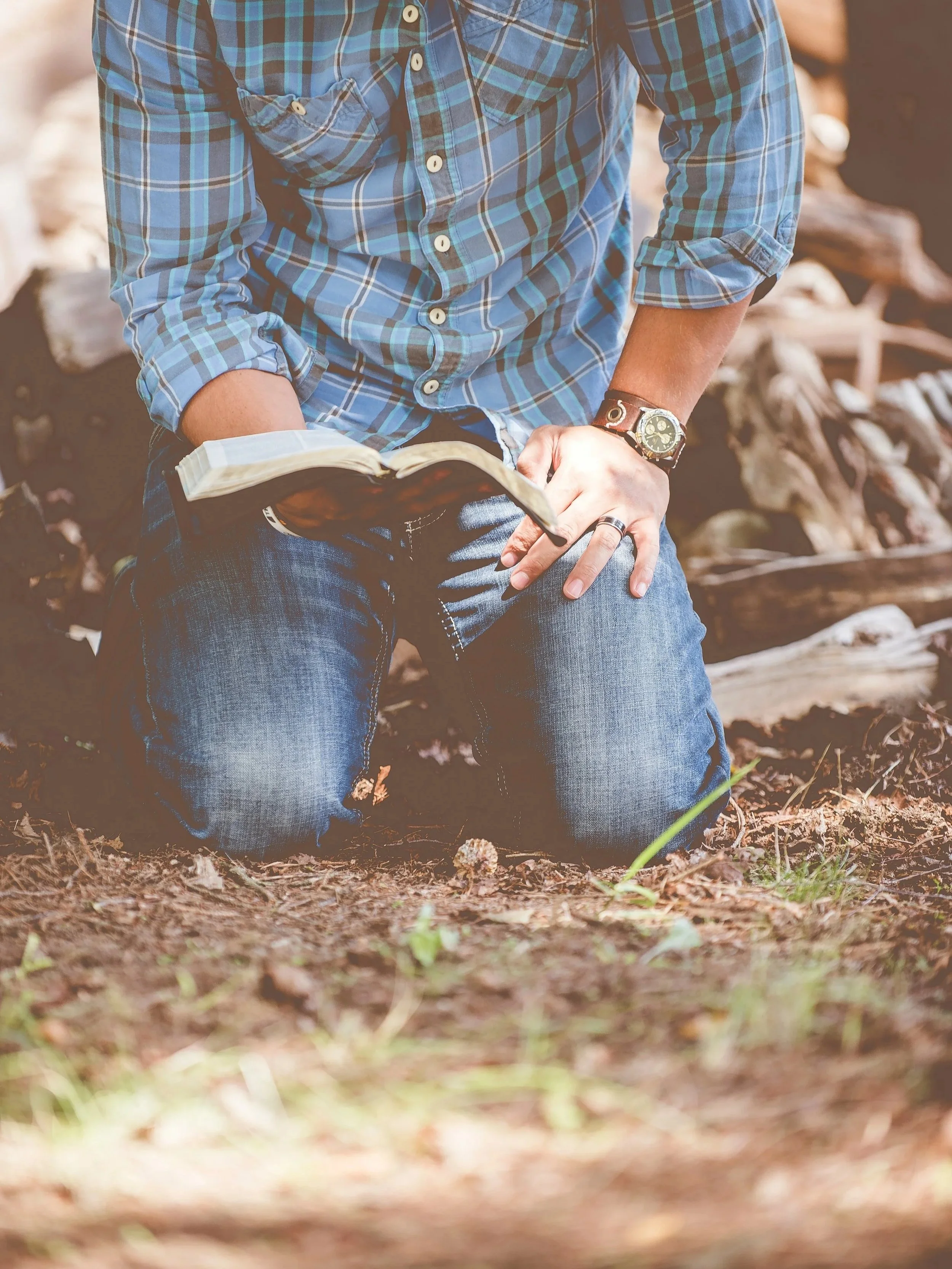 Person kneeling outdoors on dirt ground, wearing a blue plaid shirt and jeans, holding an open Bible.