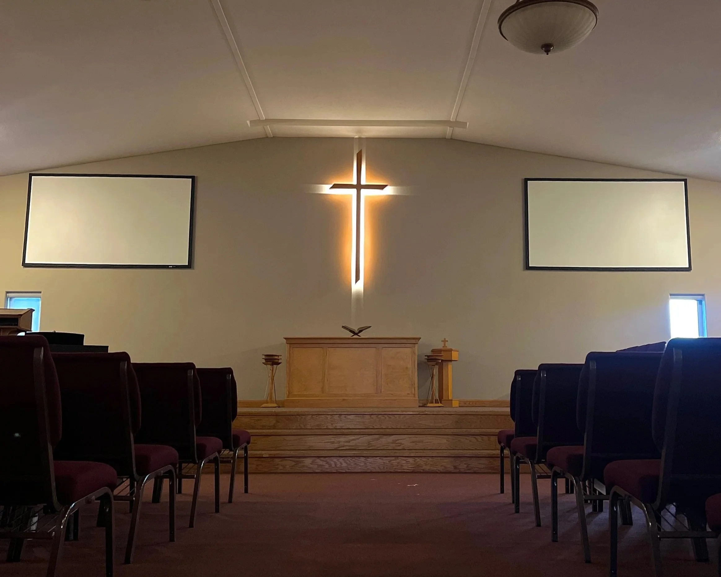 Interior of a Christian church sanctuary with a wooden altar, a glowing cross on the wall behind, two blank screens on each side, and rows of chairs facing the altar.
