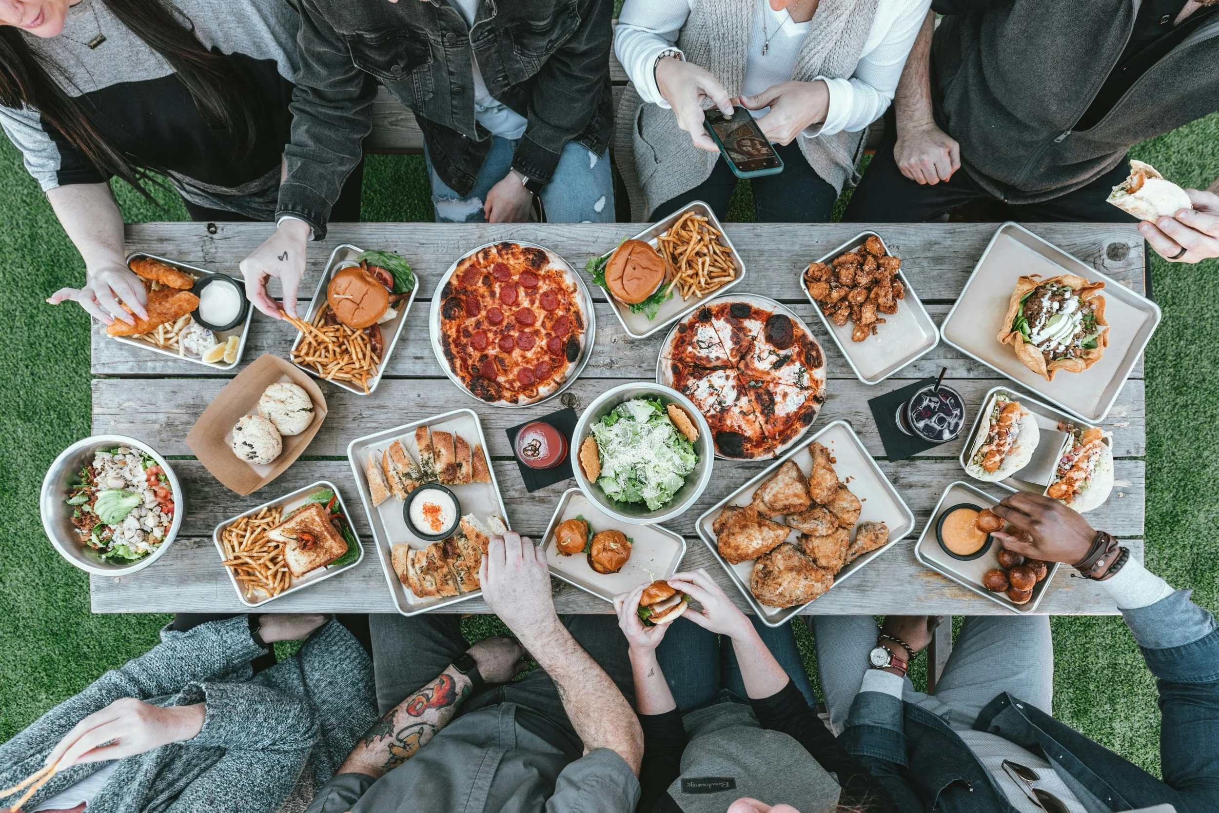 A group of people enjoying a meal at an outdoor wooden table filled with various dishes like pizza, salads, sandwiches, fried chicken, and drinks.