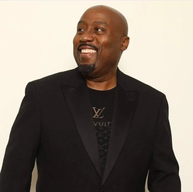 A smiling African American man with a goatee, wearing a black blazer and Louis Vuitton t-shirt, standing against a plain light-colored background.