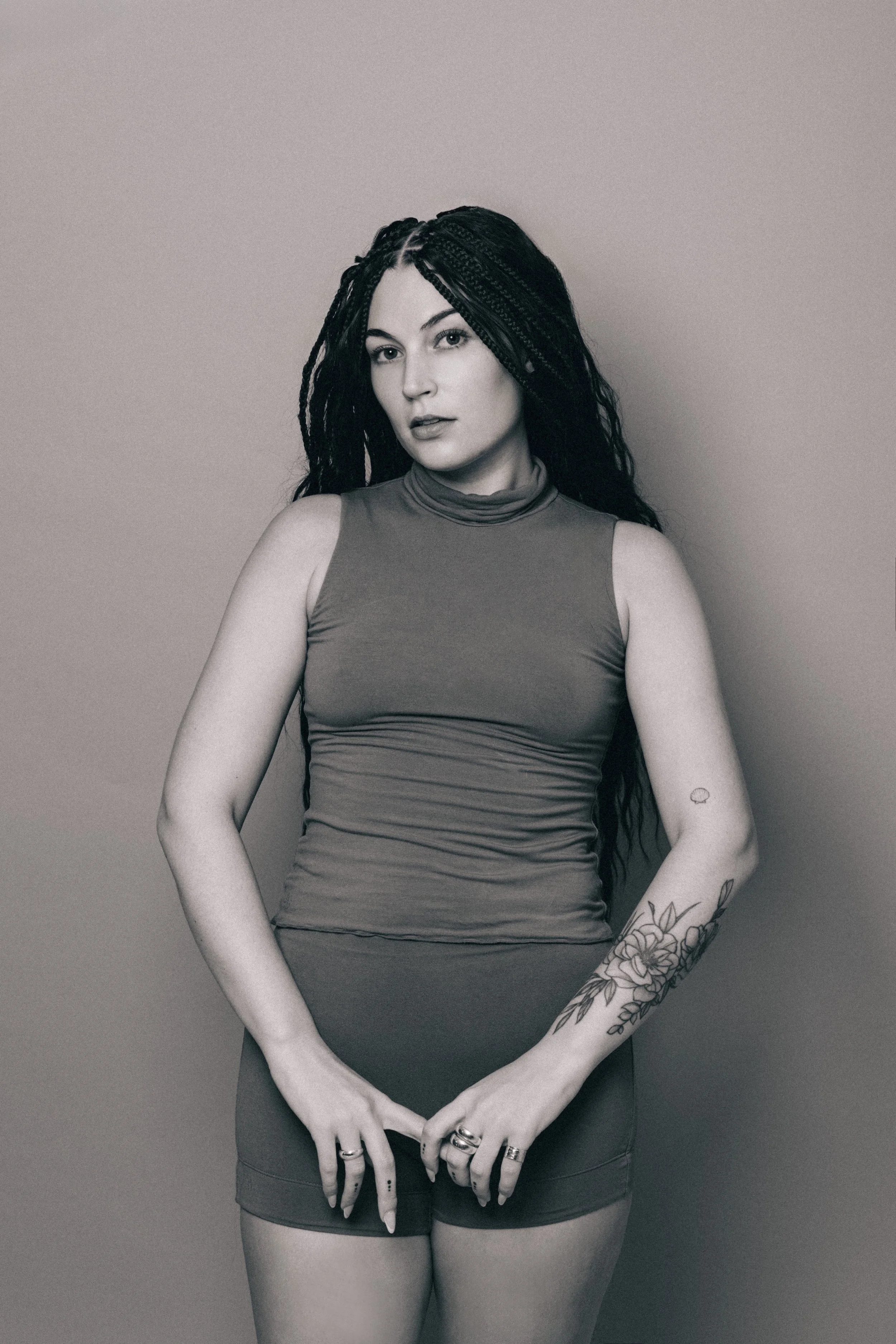 Black-and-white photo of a woman with long, styled hair, wearing a sleeveless turtleneck top and shorts, standing against a plain background, with tattoos on her arms.