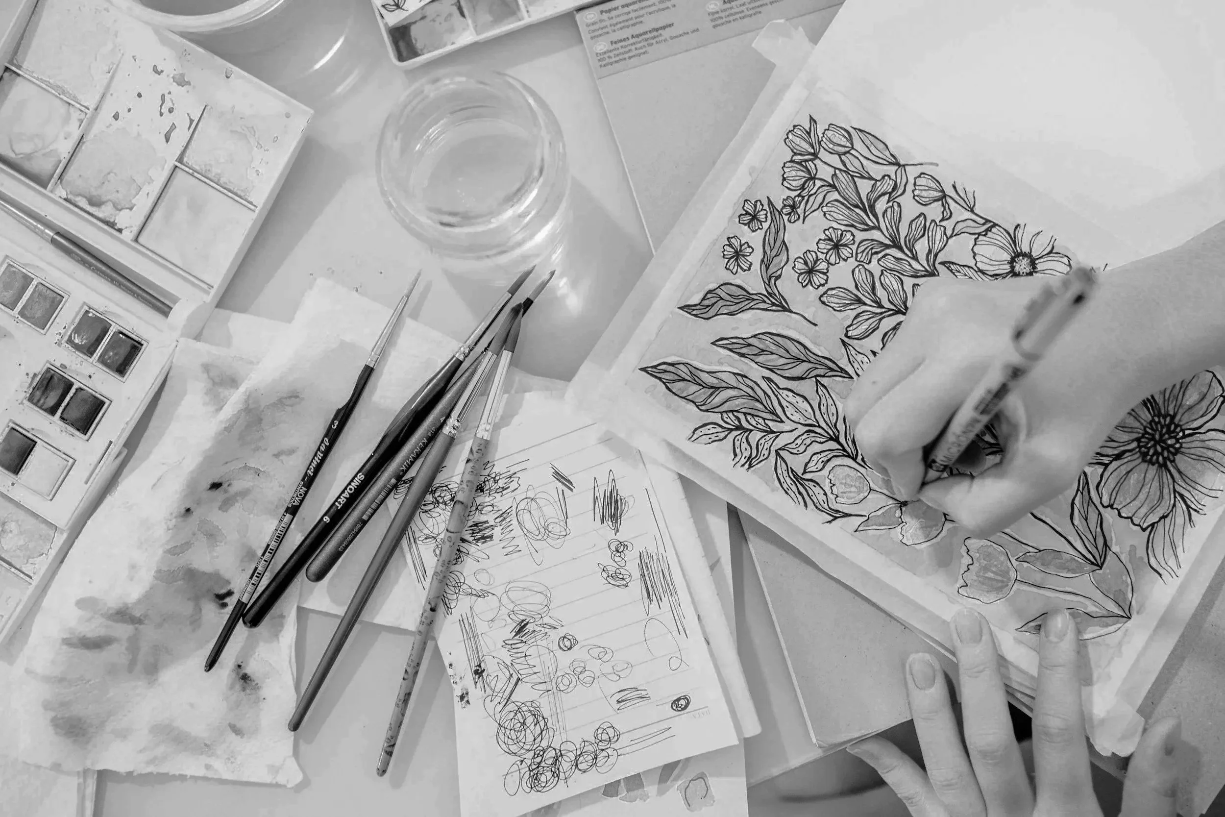 A person is coloring a detailed floral design on paper using a marker. The workspace includes watercolor paints, brushes, a glass of water, and drawing sketches.