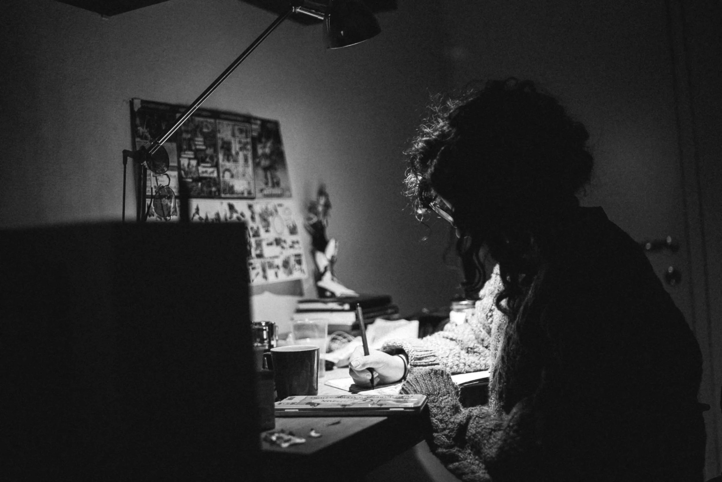 A person with curly hair and glasses drawing at an art table. There are books, a mug, and various items on the desk, with pictures and posters on the wall behind.