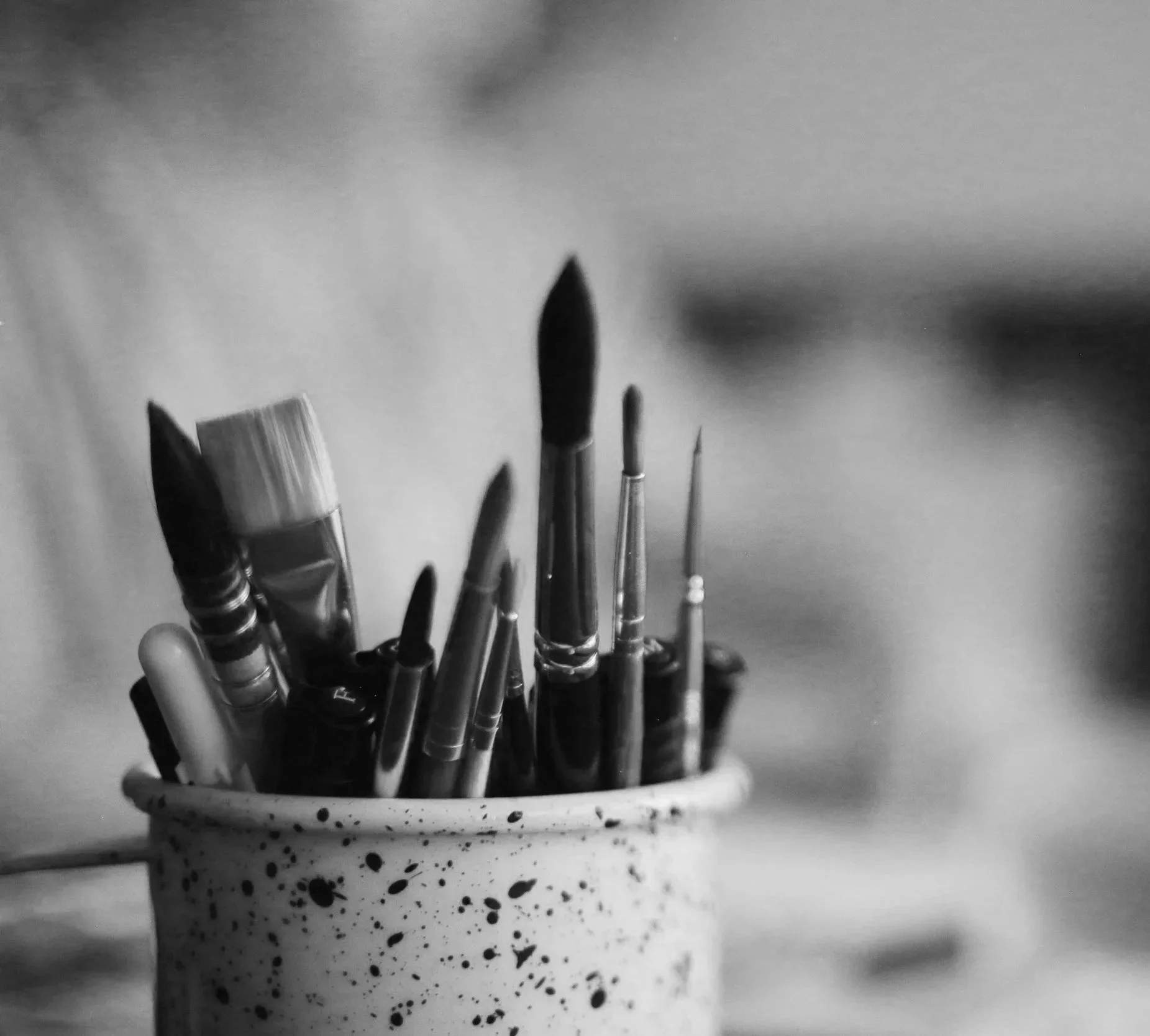 A ceramic mug with black paint splatters filled with various paintbrushes and art tools, seen in black and white.