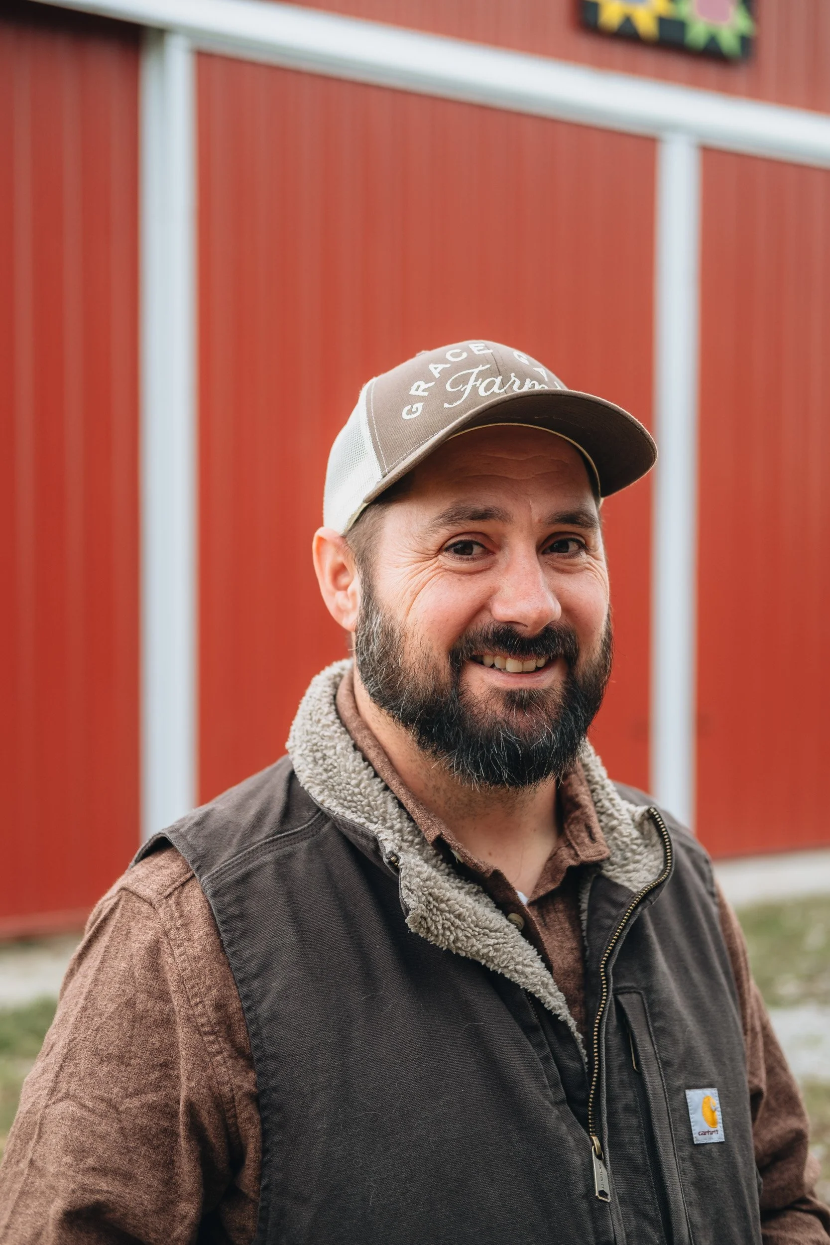 A smiling man with a beard and mustache wearing a tan cap, a brown shirt, and a black Carhartt vest standing in front of a red barn with white trim.
