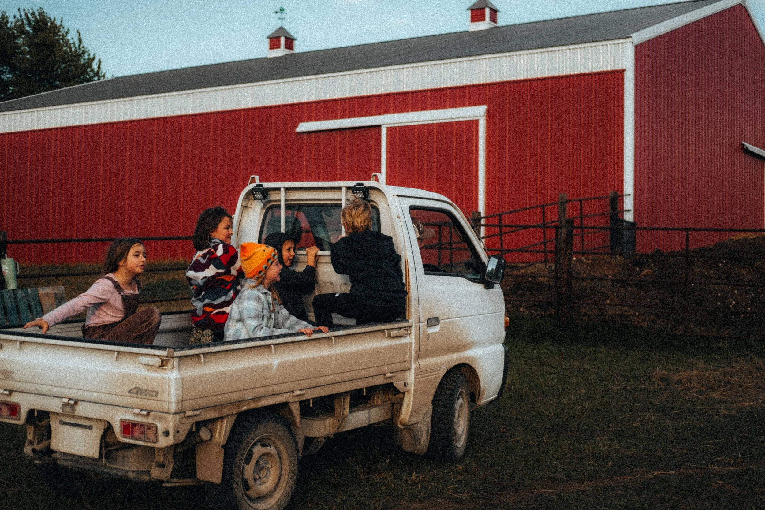 A small white pickup truck parked on grass with five children sitting in the truck bed, facing a red barn, during sunset.
