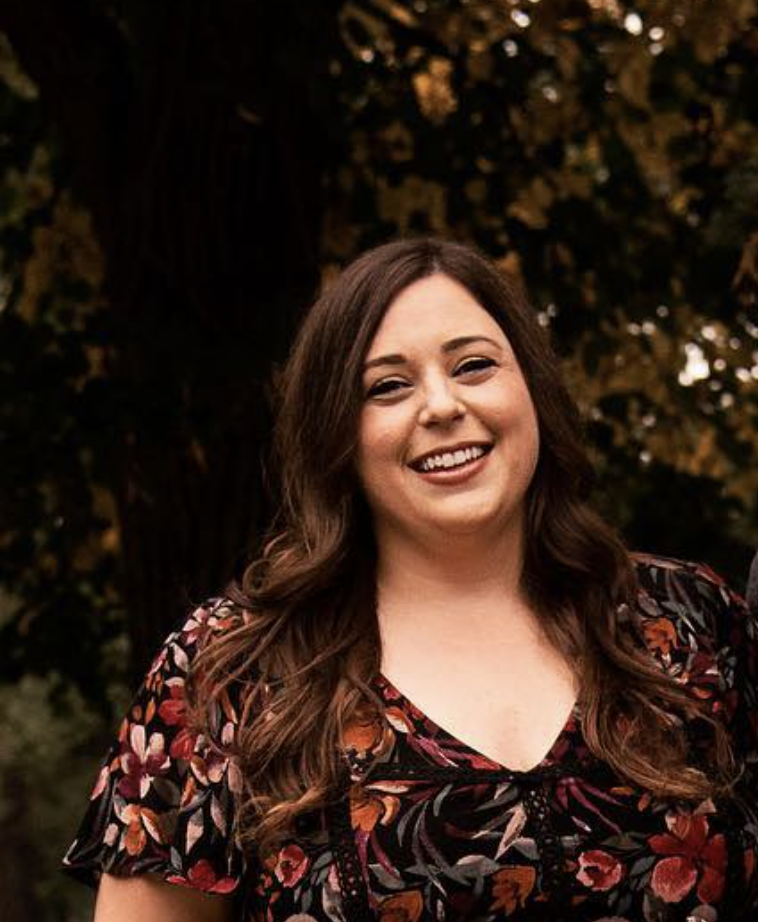 A woman with long brown hair smiling outdoors at night, wearing a black floral dress with red, orange, and pink flowers, in front of a tree with autumn leaves.