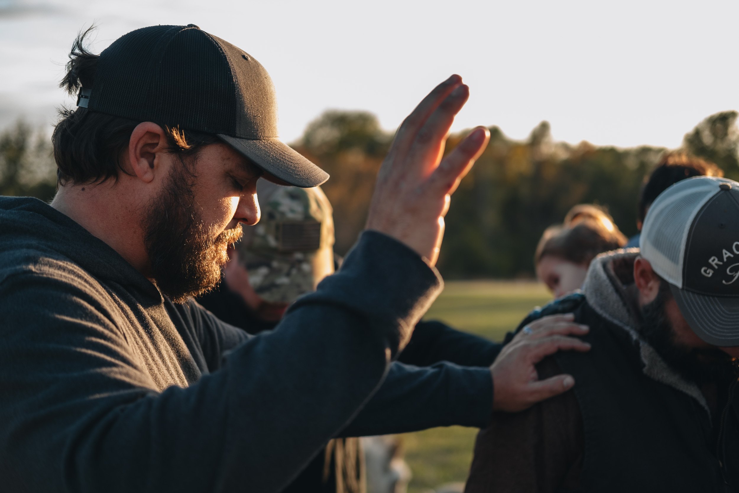 A group of people, including a man wearing a gray baseball cap, participate in a prayer or blessing outdoors at sunset, with their eyes closed.