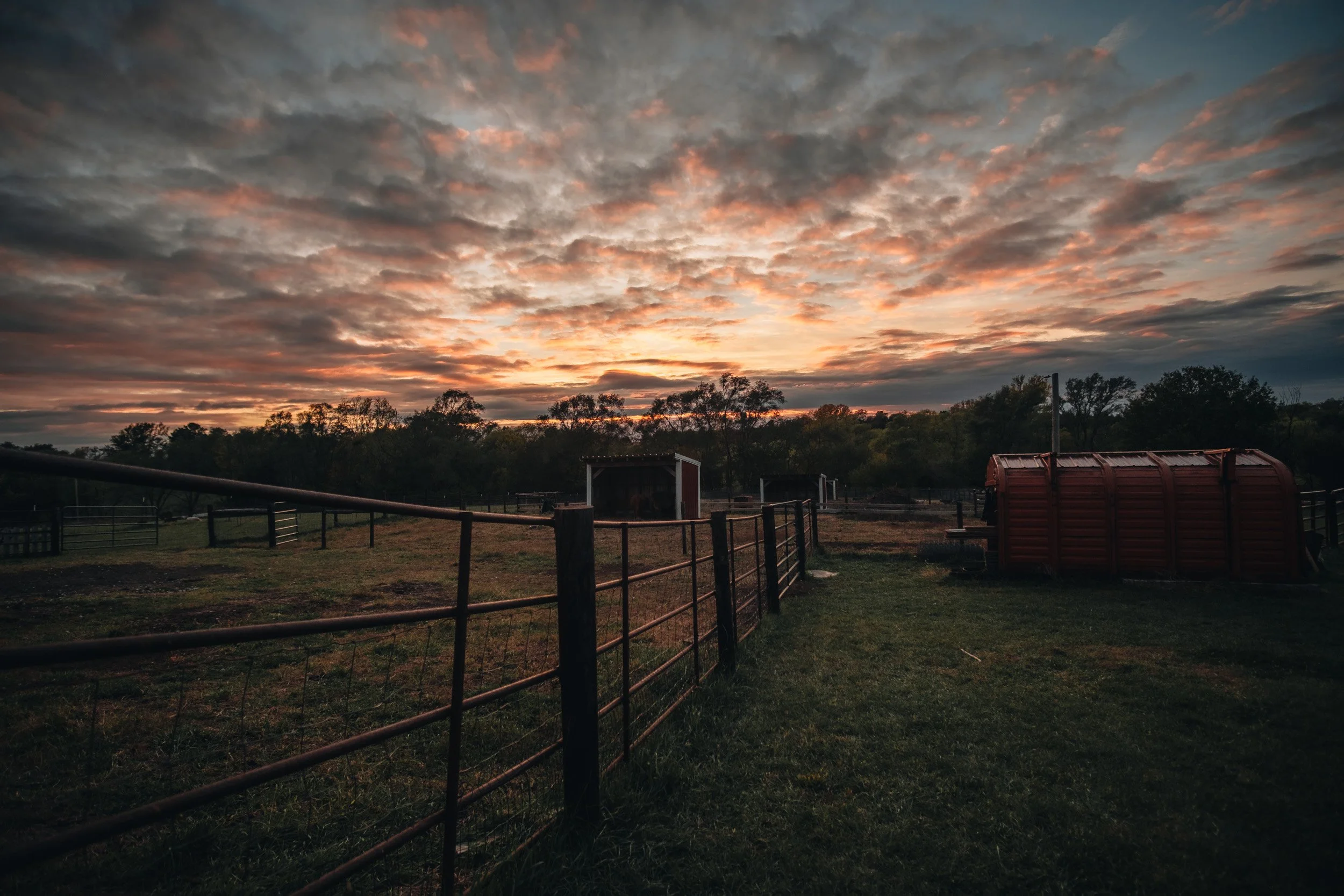 Fenced area with a small barn and a red covered water tank under a dramatic sunset sky with scattered clouds.