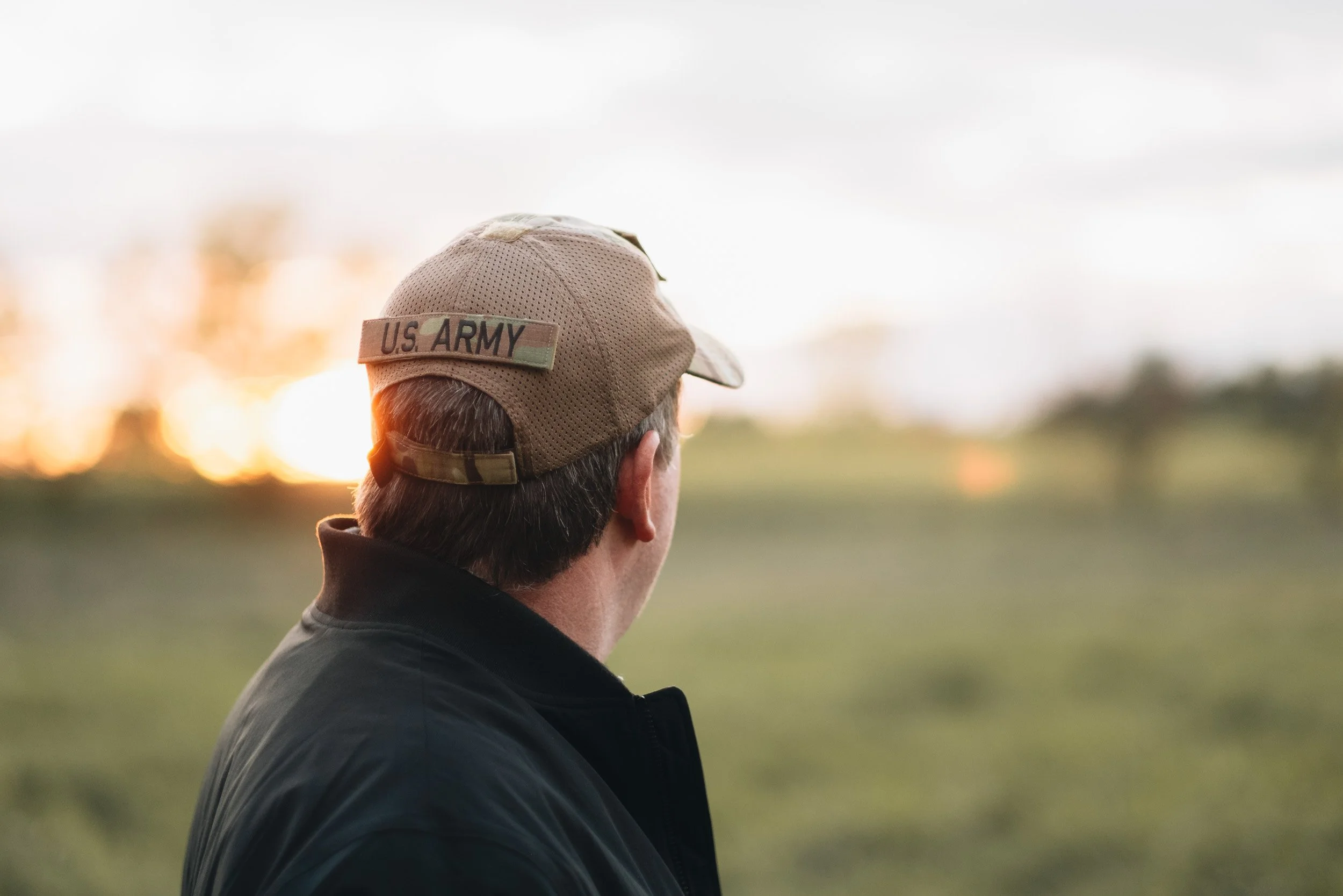 A person wearing a tan U.S. Army baseball cap and a black jacket, looking towards a sunset or sunrise on an open field.