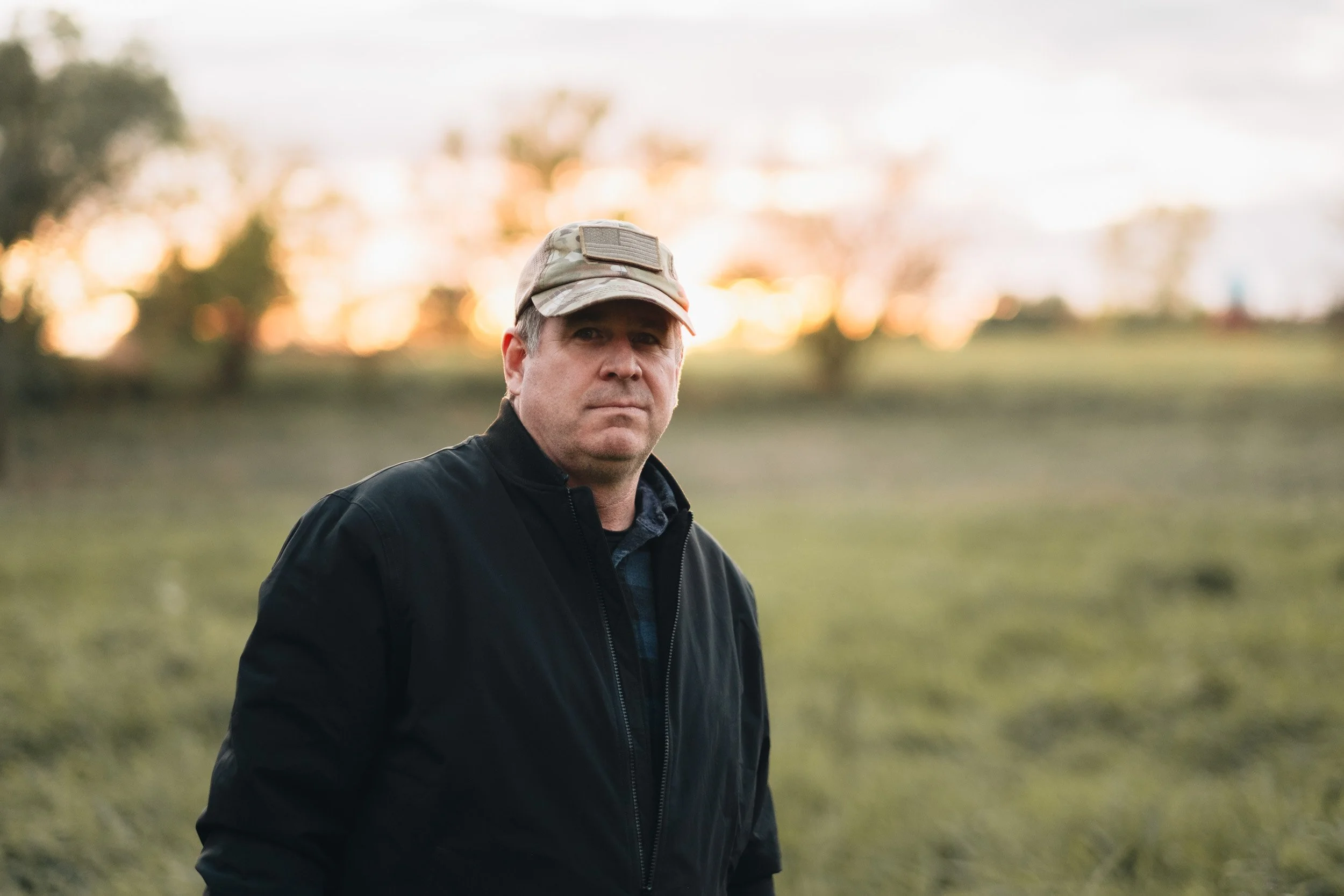 A man in a black jacket and camouflage cap stands outdoors during sunset, with a blurred background of trees and an open field.