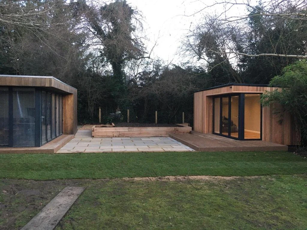 Modern backyard with two small wooden structures featuring black-framed glass doors, a paved patio area, and a lawn with some patches of grass and a wooden plank path, surrounded by trees.