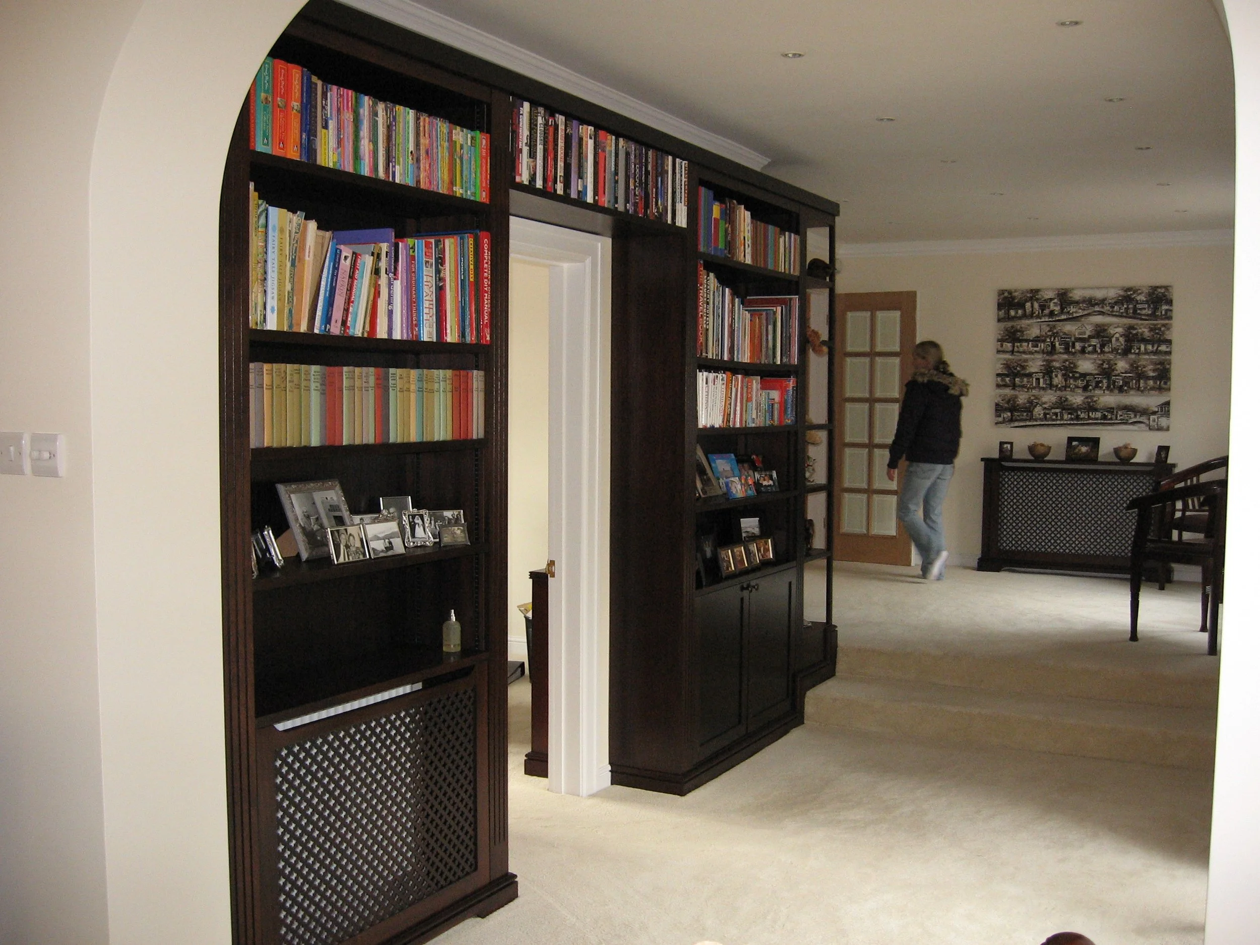 A living room with dark wood bookshelves filled with colorful books and family photographs, a beige carpeted floor, a white wall, and a person walking toward a door with glass panels.