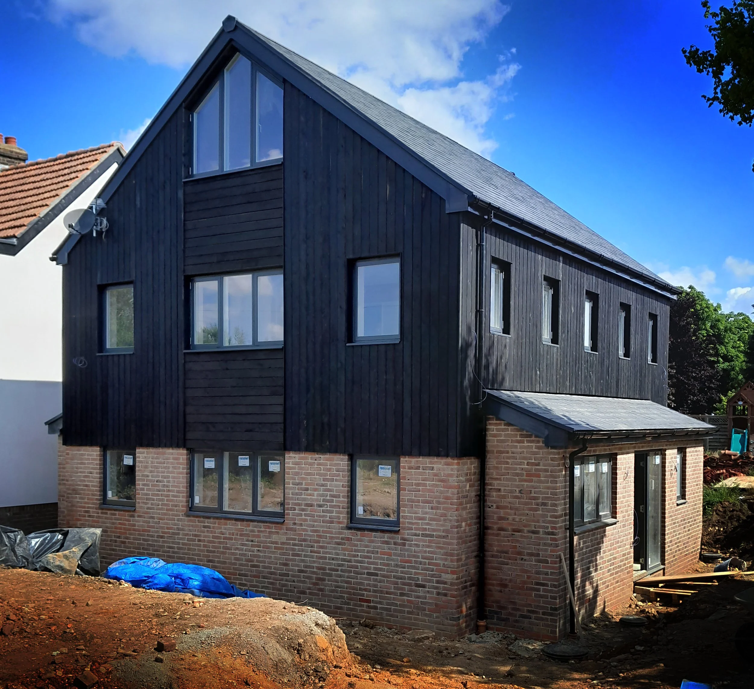 A modern house under construction with a brick foundation and black wood siding, multiple windows, and a sloped roof.