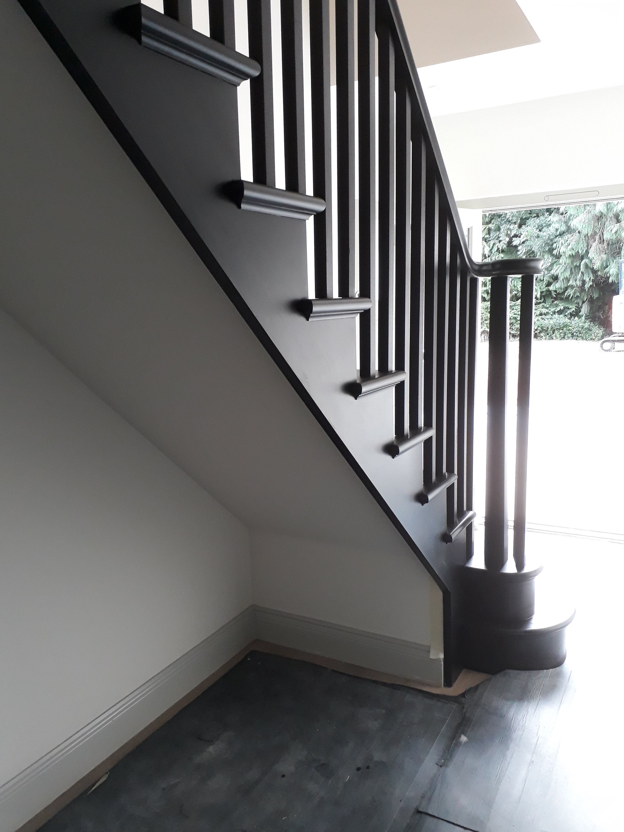Black wooden staircase with vertical balusters and rounded baseboard at bottom, leading upstairs in a house with white walls and hardwood flooring