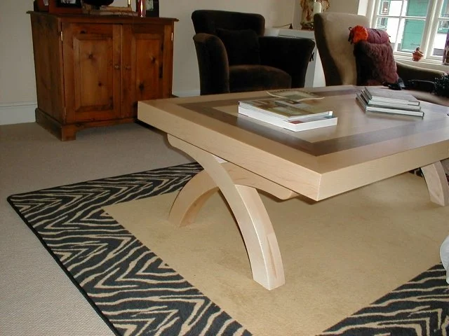A living room with a wooden sideboard, two armchairs, a window with plants, and a rectangular coffee table with magazines, on a patterned rug.