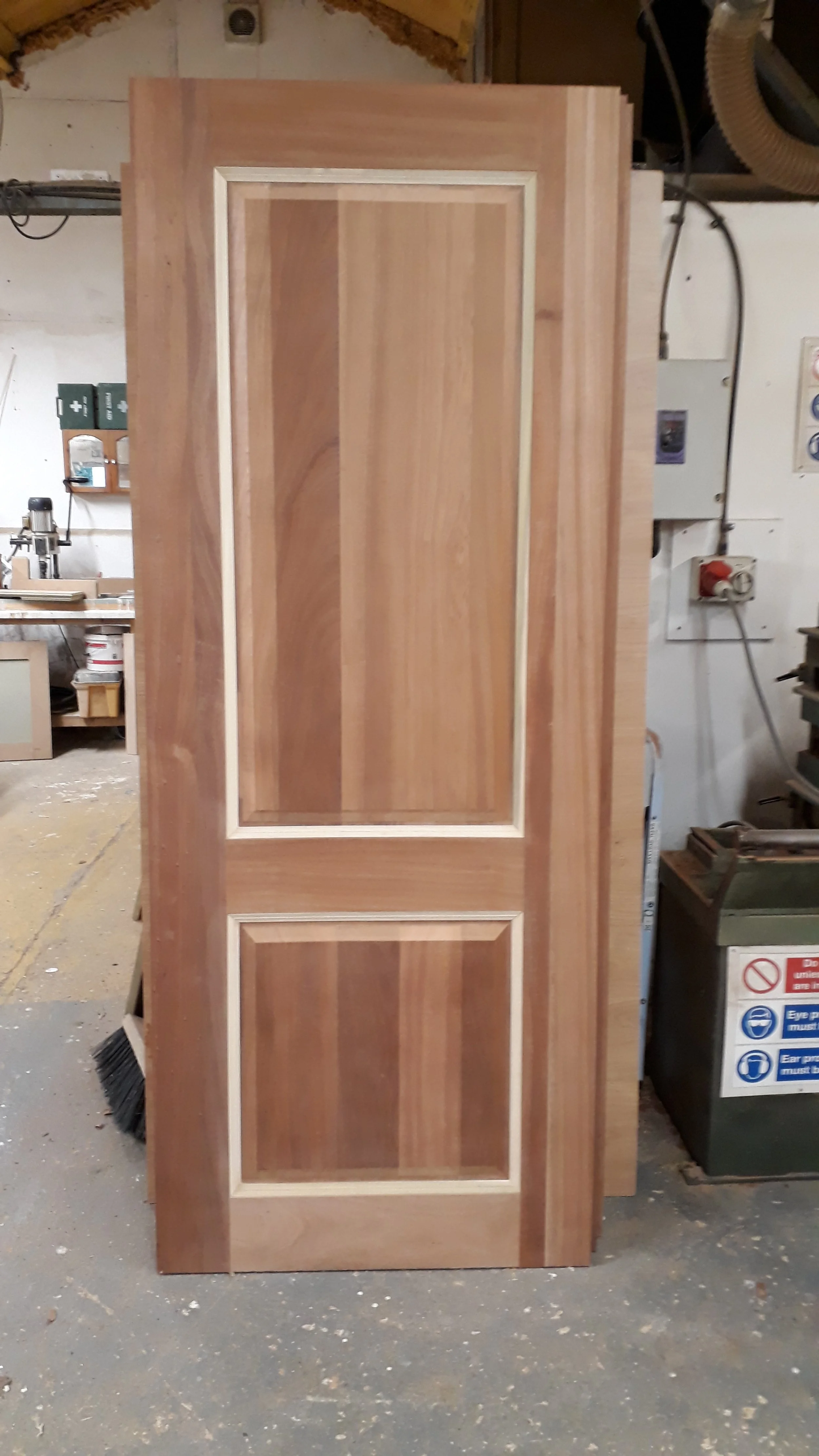 A wooden cabinet door with two rectangular panels, made of natural wood with a light-colored trim, standing upright in a woodworking shop.