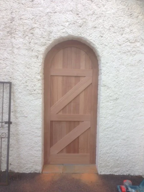 A small wooden door with an arched top set into a white, textured stucco wall.