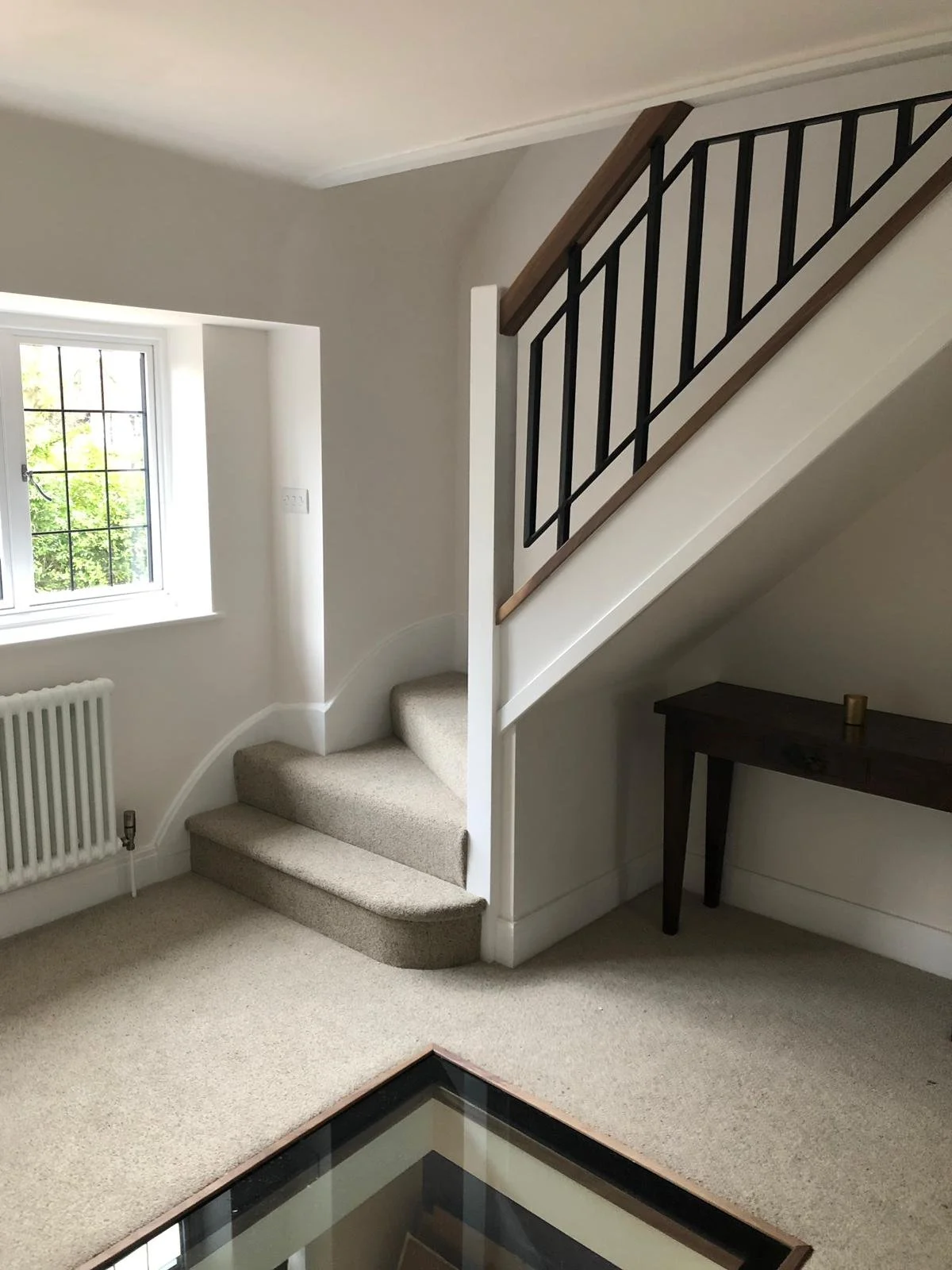 Interior of a room with beige carpet, a staircase with beige carpeted steps, and a wooden and black metal railing. A window with a view of trees, a radiator, and a small dark wooden table are visible.