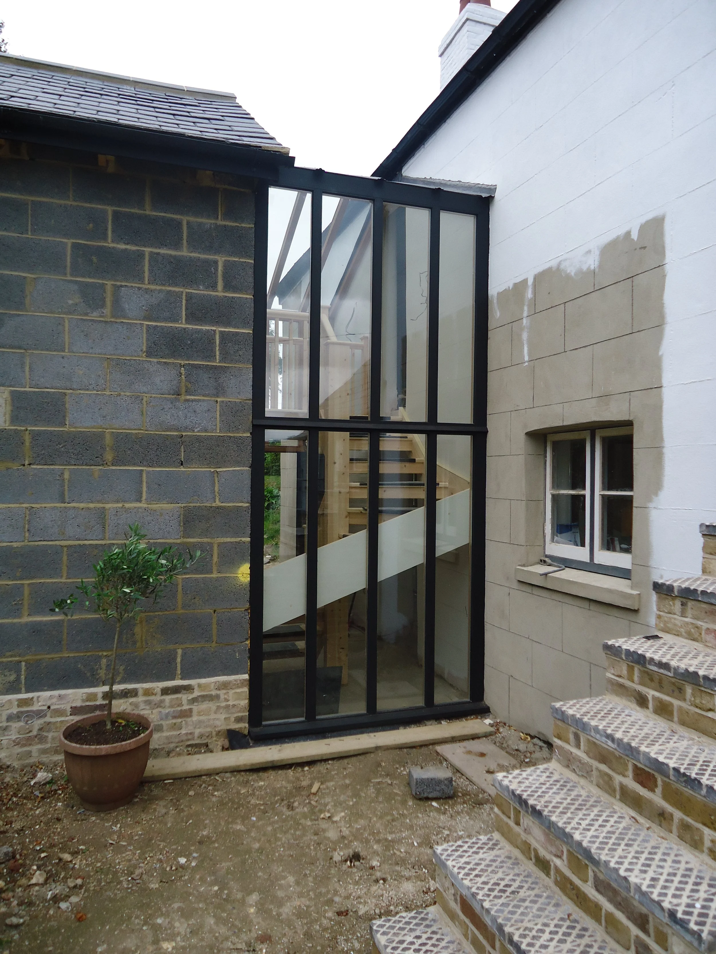 New glass and metal staircase enclosure between two houses, with brick steps, a small window, and a potted plant outside.