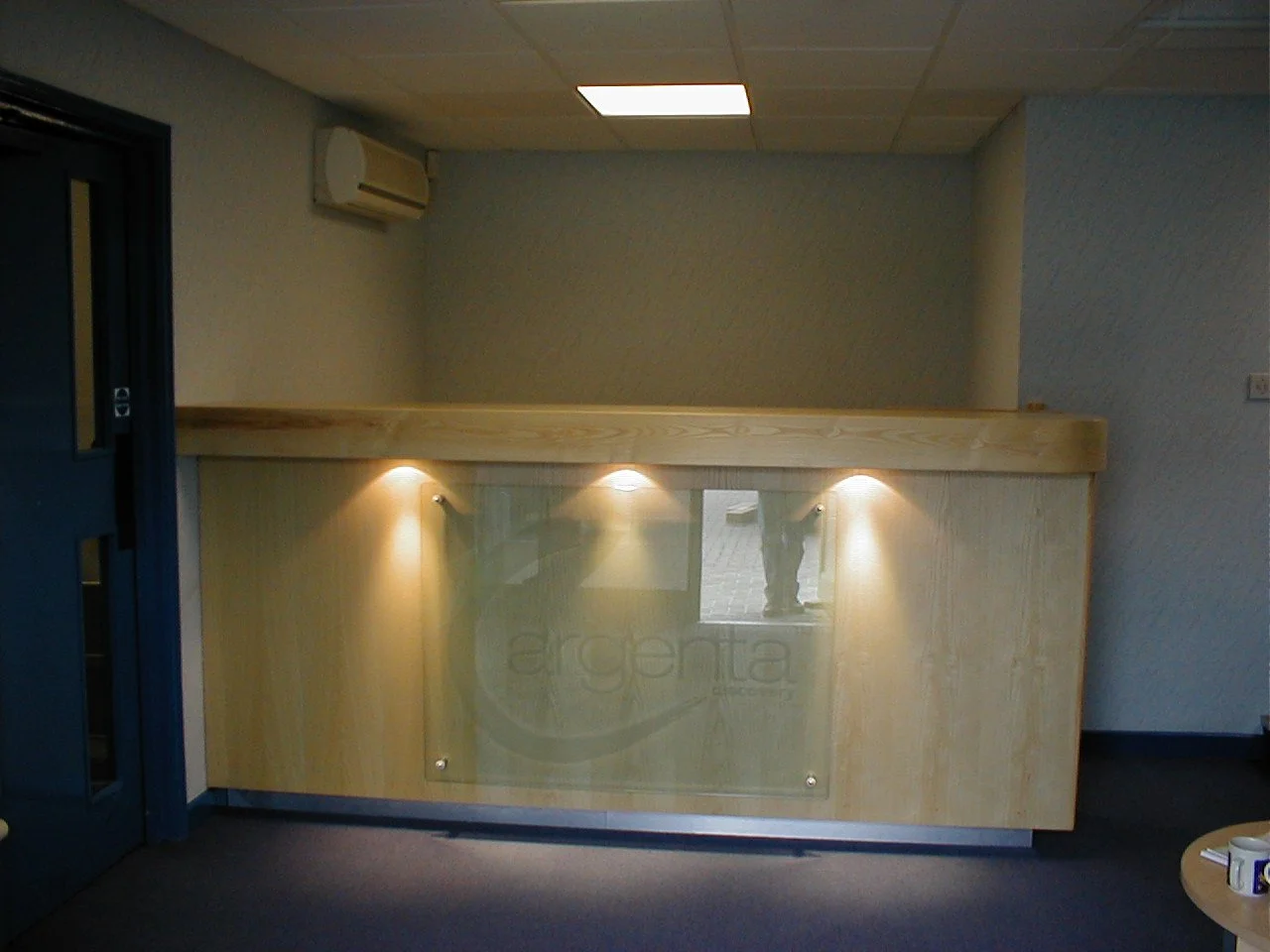 Wooden reception desk with glass panel and the word 'argenta' engraved, illuminated by three small spotlights, in a room with gray walls, ceiling with a light fixture, and an air conditioning unit.