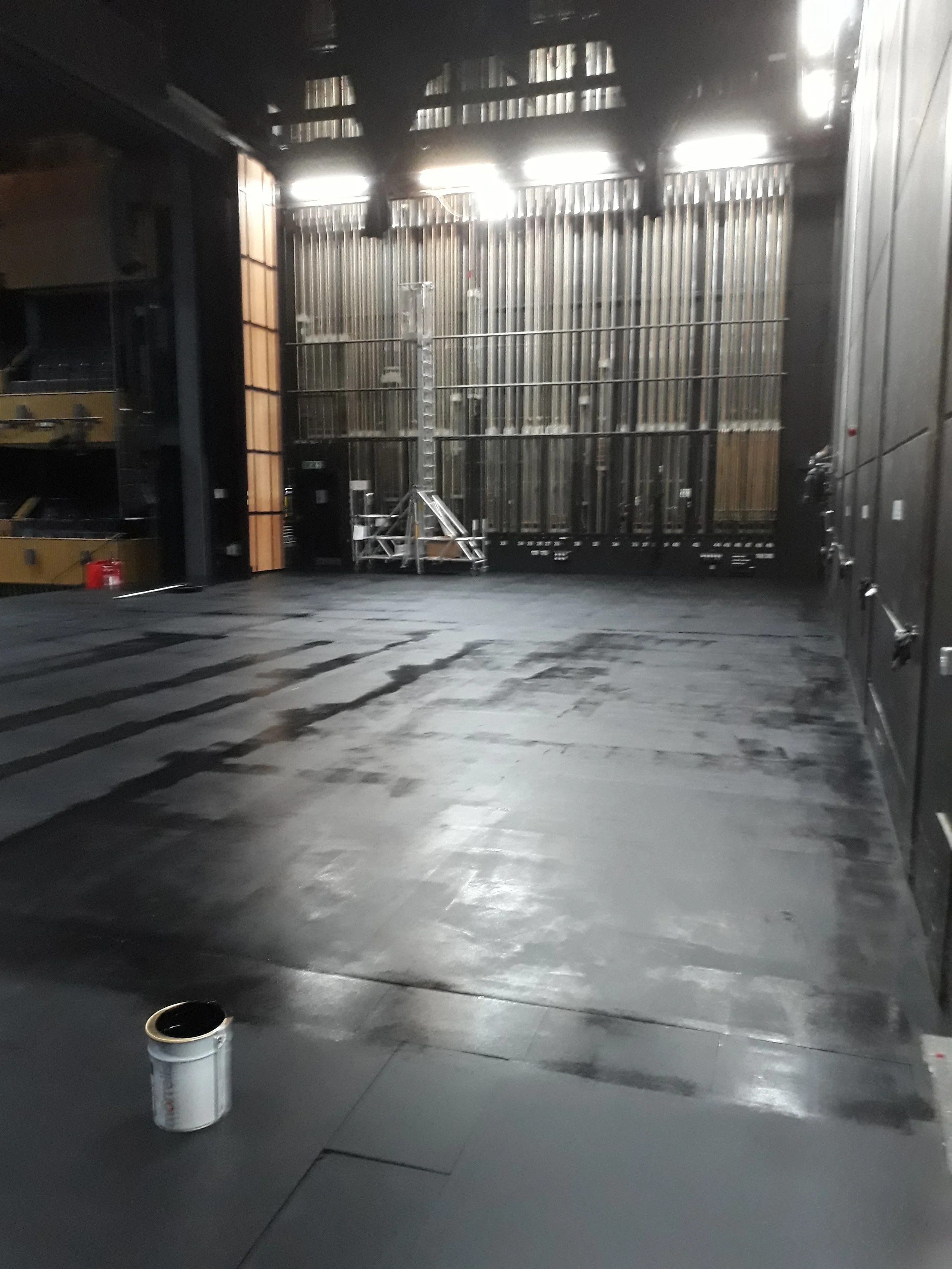 Stage area with partially painted black flooring, a paint can in the foreground, and stage infrastructure such as vertical metal pipes and wooden structures at the back.