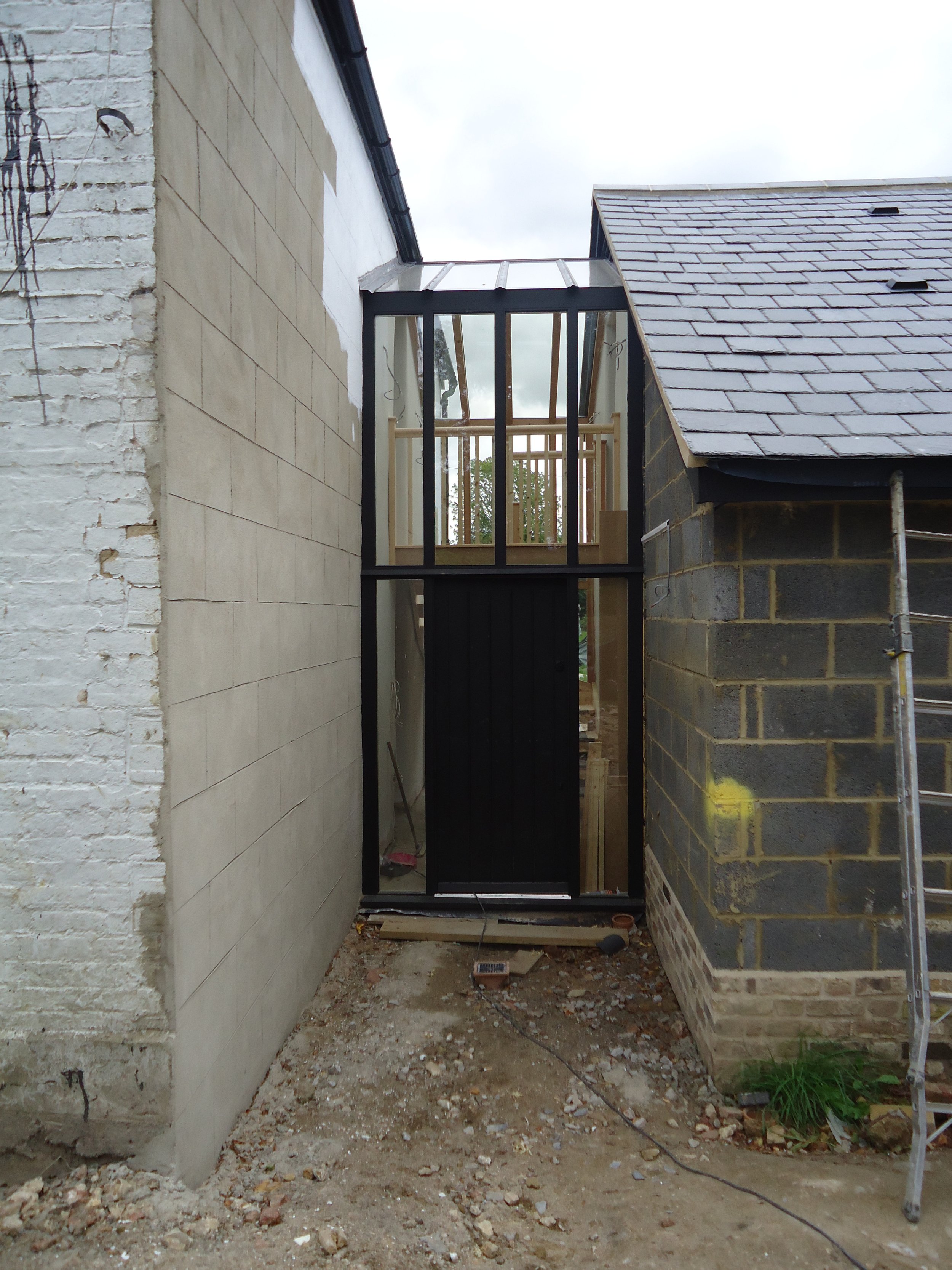 Construction site showing the frame of a new glass extension or door between two existing buildings, with a temporary black door and scaffolding on the right.