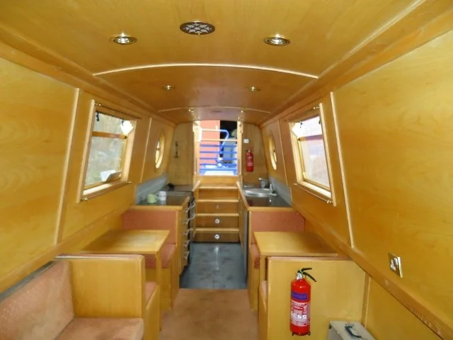 Interior of a small wooden camper or boat with built-in seating, windows, a small kitchen with a sink, and a fire extinguisher.